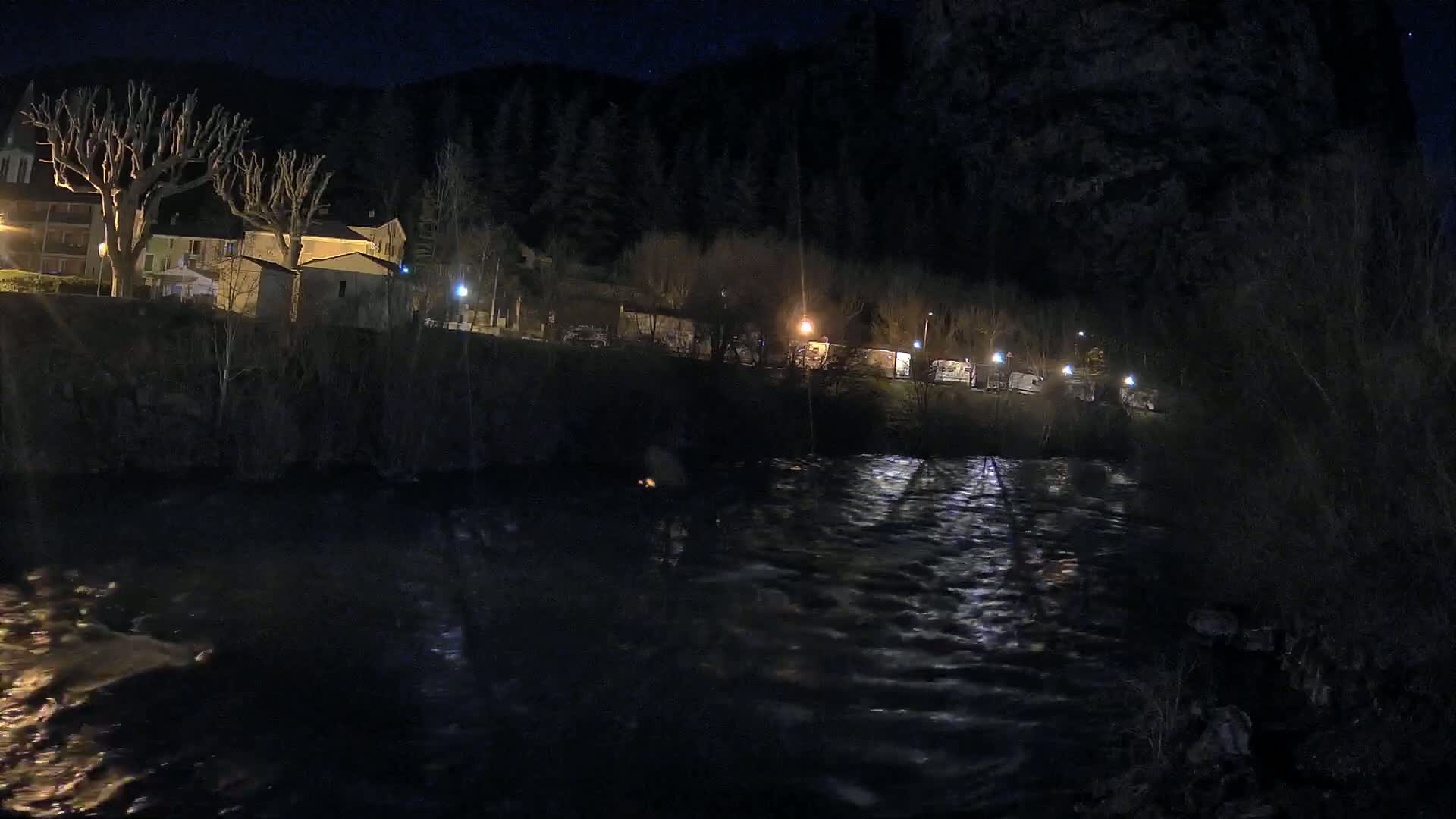 A nighttime view of a flowing river with a small village on a hillside under a starry sky.