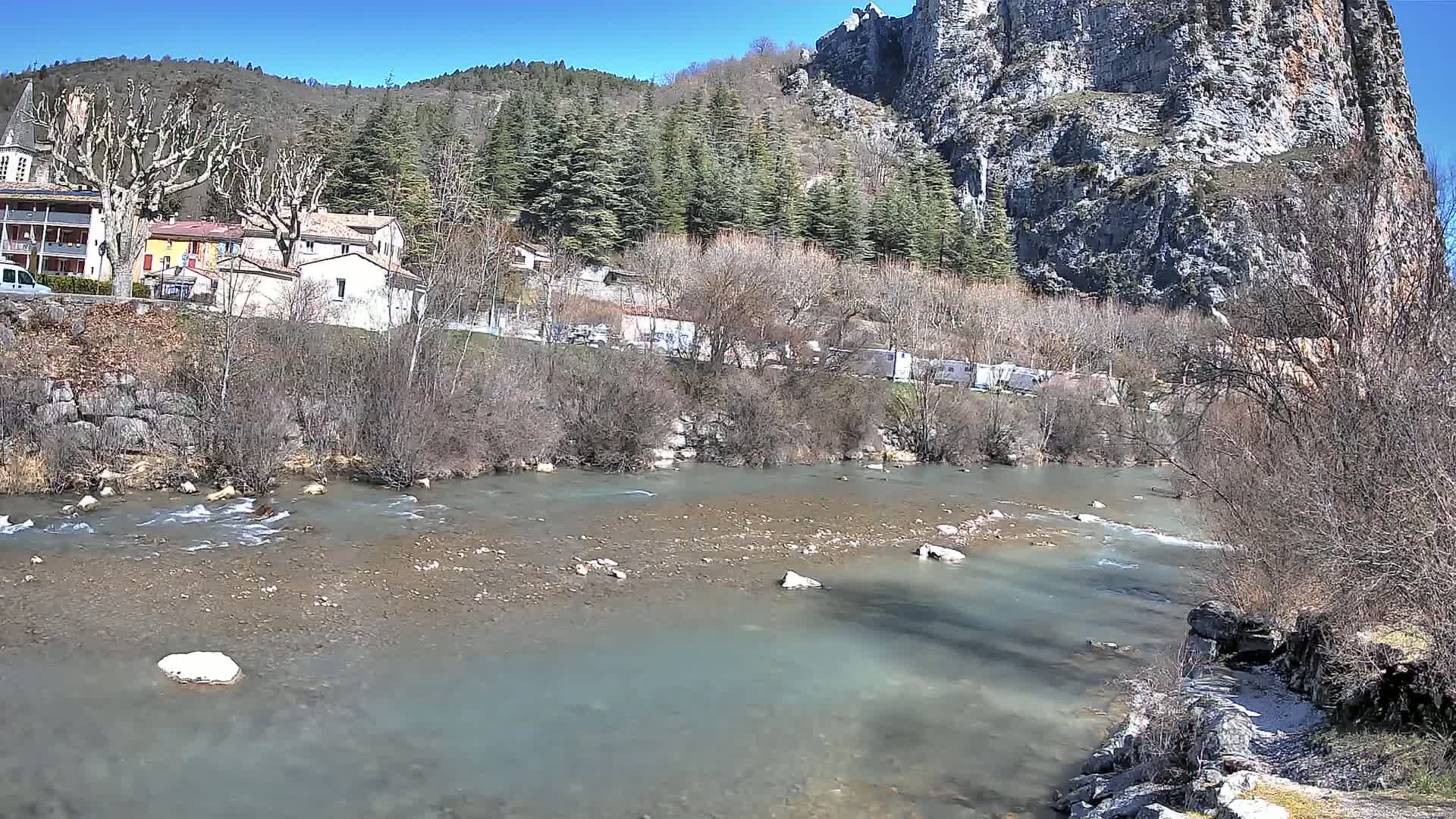 A river flows gently past a small village nestled at the base of a large, rocky cliff on a sunny day.