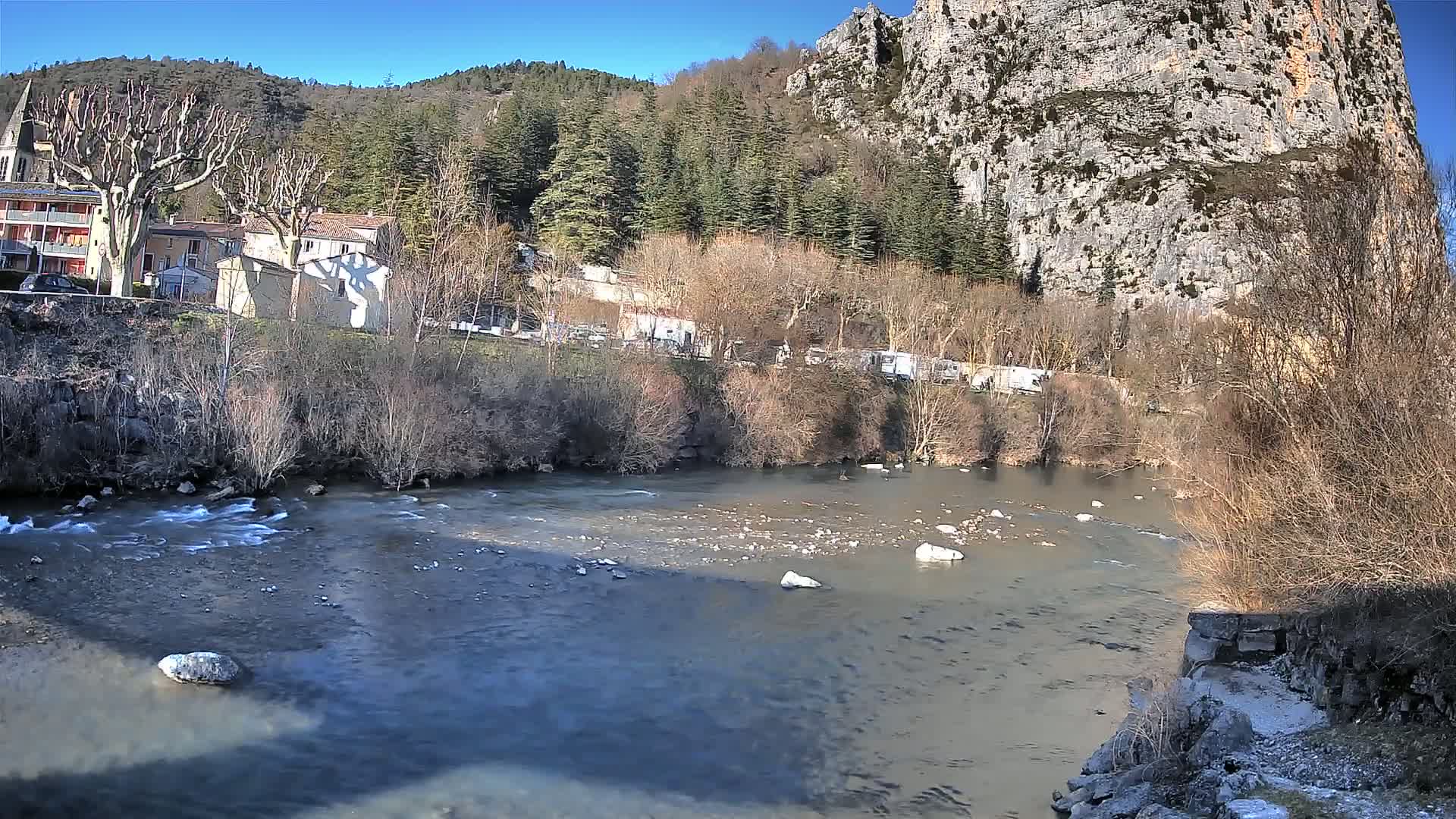 A river flows past a village nestled at the base of a large, rocky cliff under a clear blue sky.