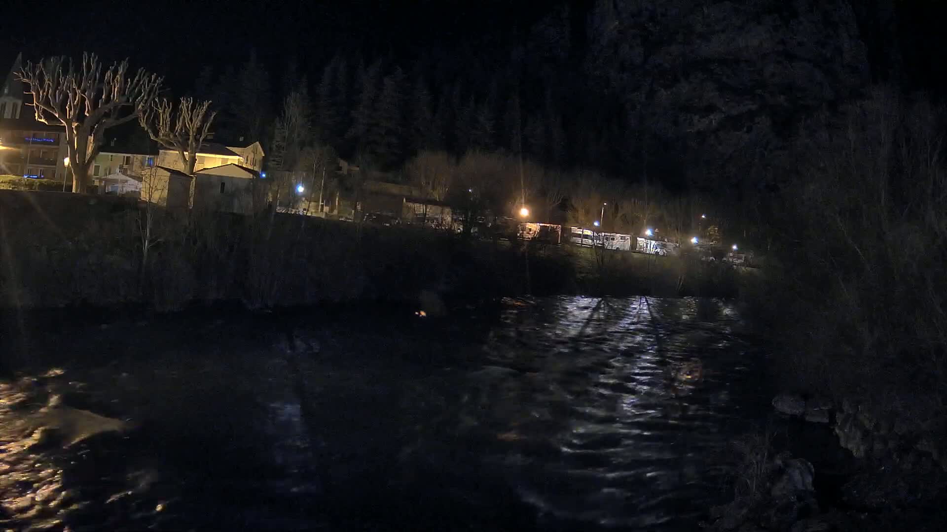 A dark, nighttime scene shows a flowing river reflecting lights from buildings and vehicles along its bank, under a mostly clear sky.