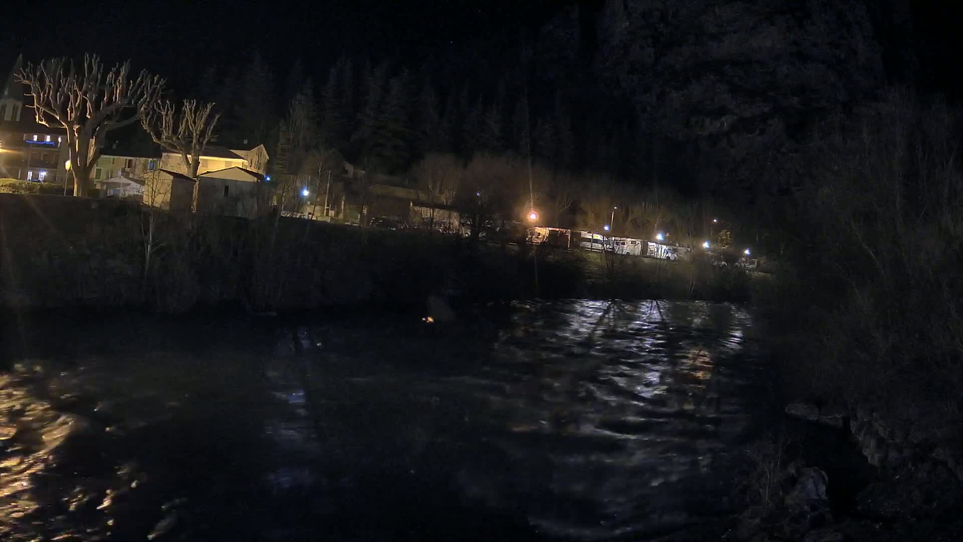 A nighttime view of a river flowing past a village with sparsely lit buildings and trees under a dark sky.