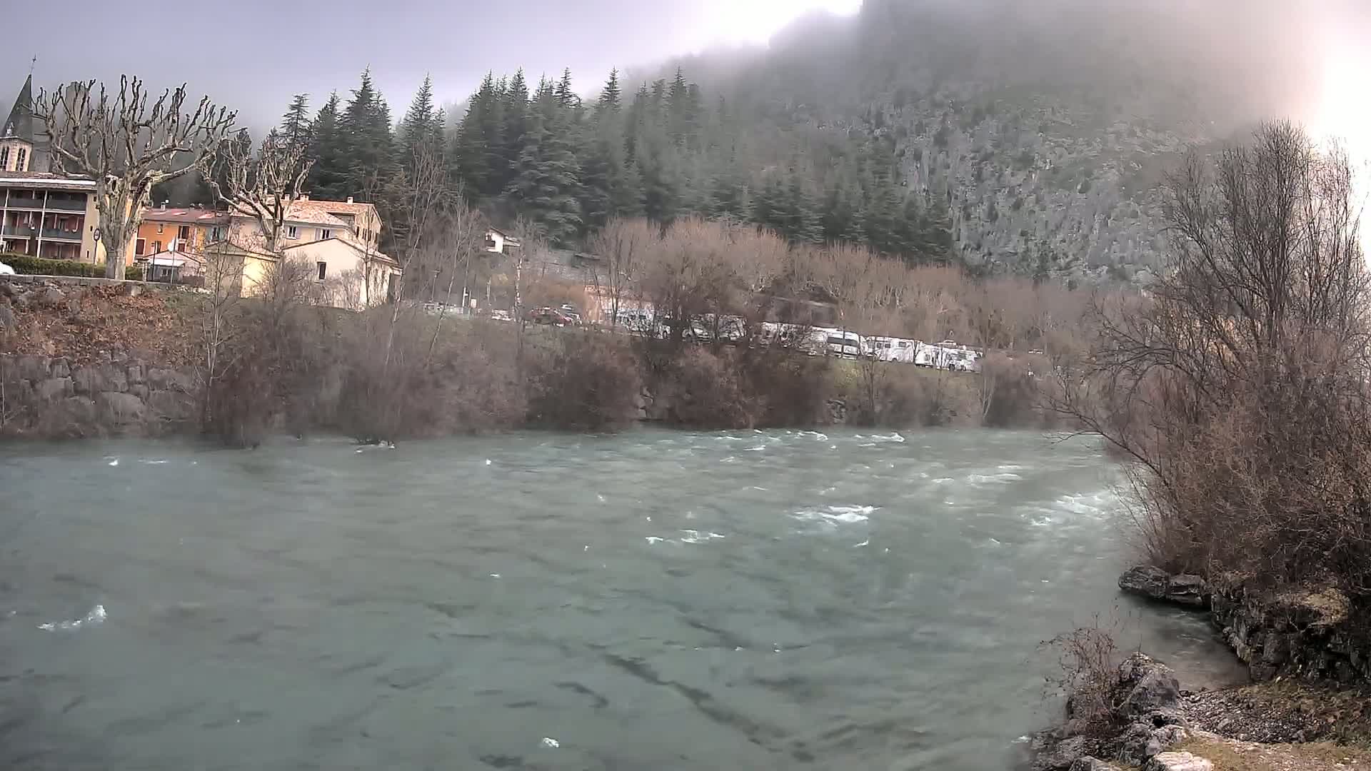 A turbulent river flows in front of a village nestled at the base of a misty, forested mountain.