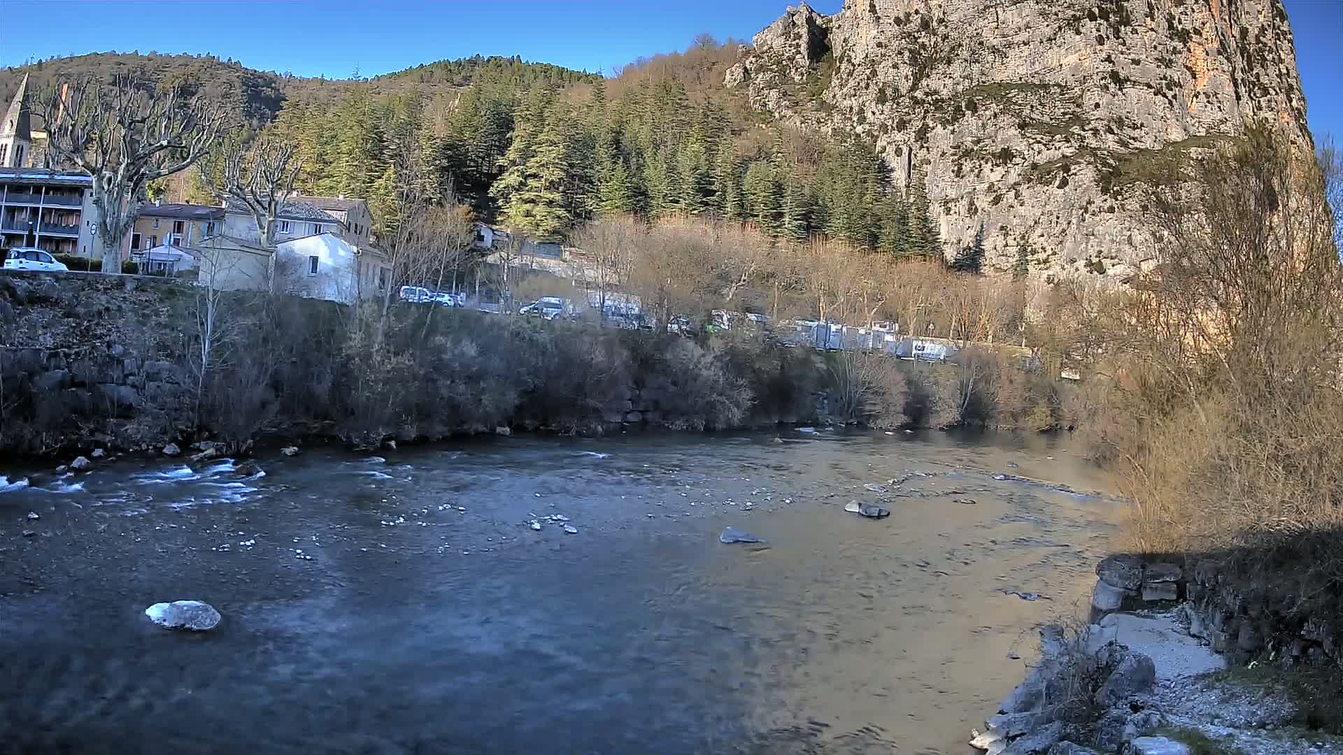 A river flows past a village nestled at the base of a steep, rocky cliff under a clear blue sky.