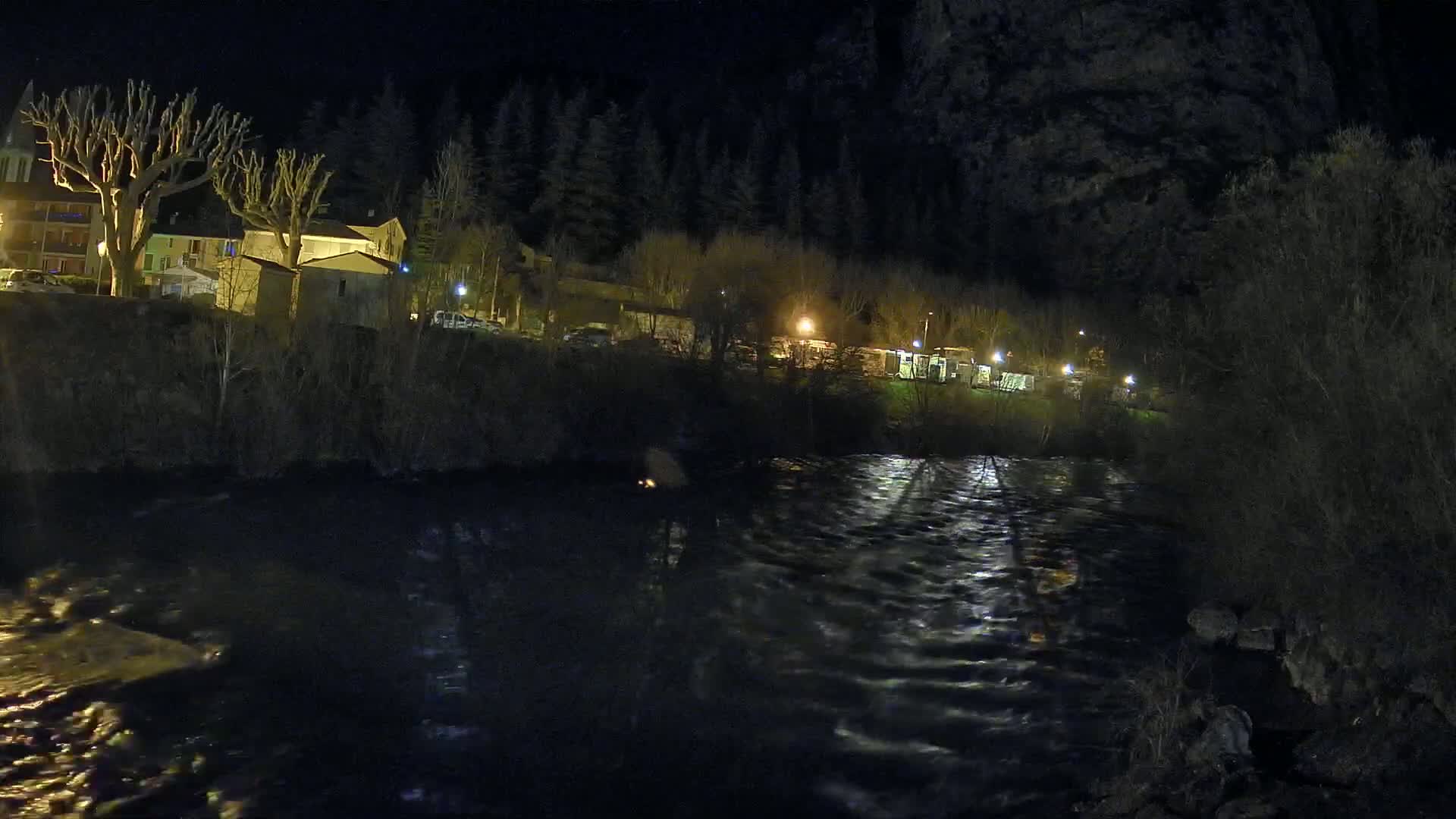 A river flows under a nighttime sky past a village of low buildings and leafless trees, illuminated by scattered streetlights.