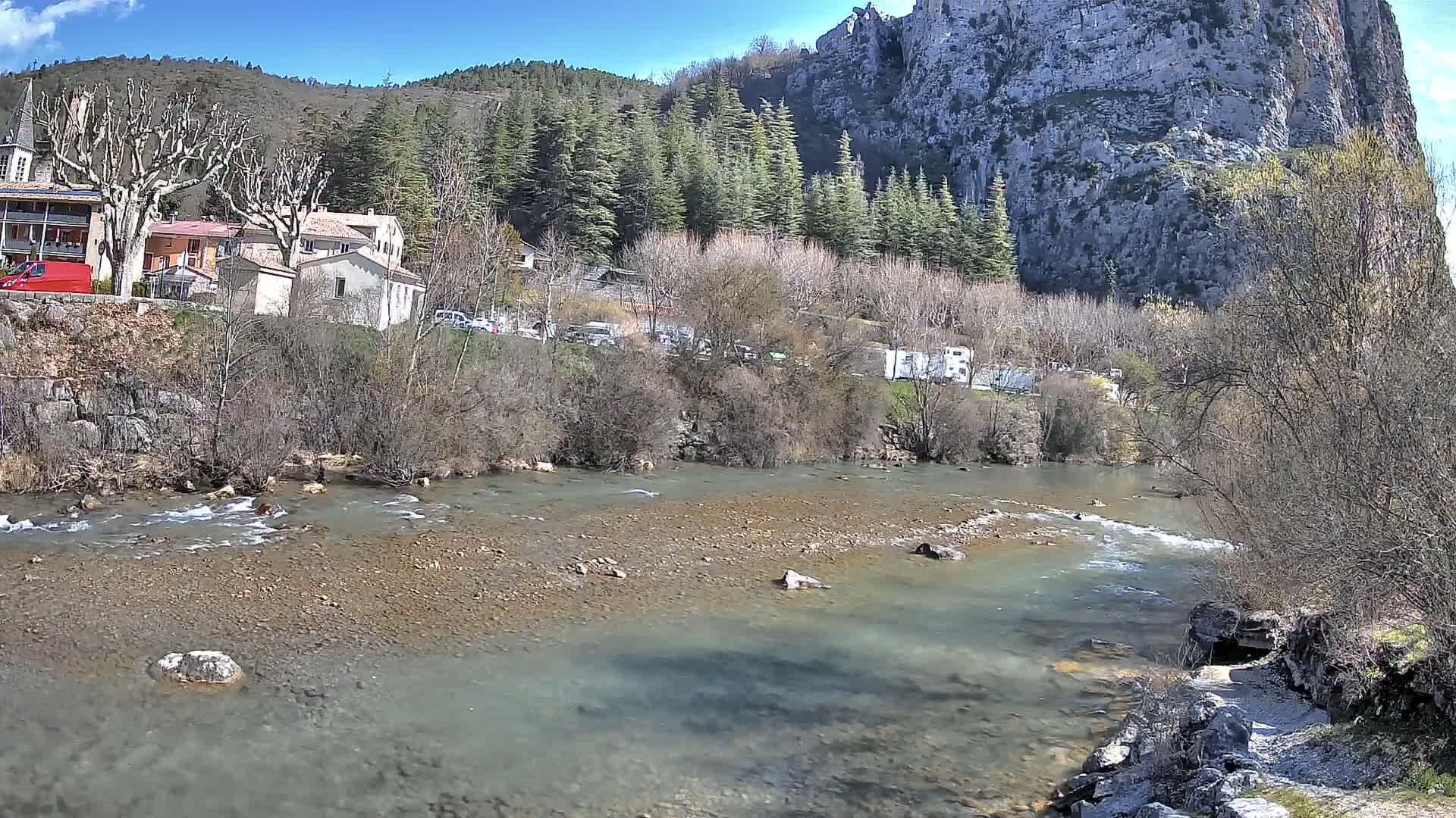A shallow, rocky river flows past a village nestled at the base of a large, rocky cliff under a sunny sky.
