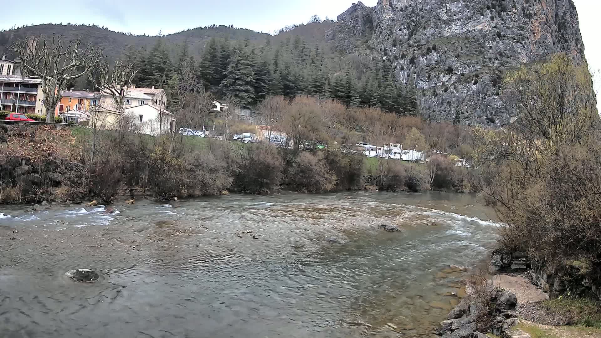 A river flows past a village nestled at the base of a steep, rocky cliff under an overcast sky.