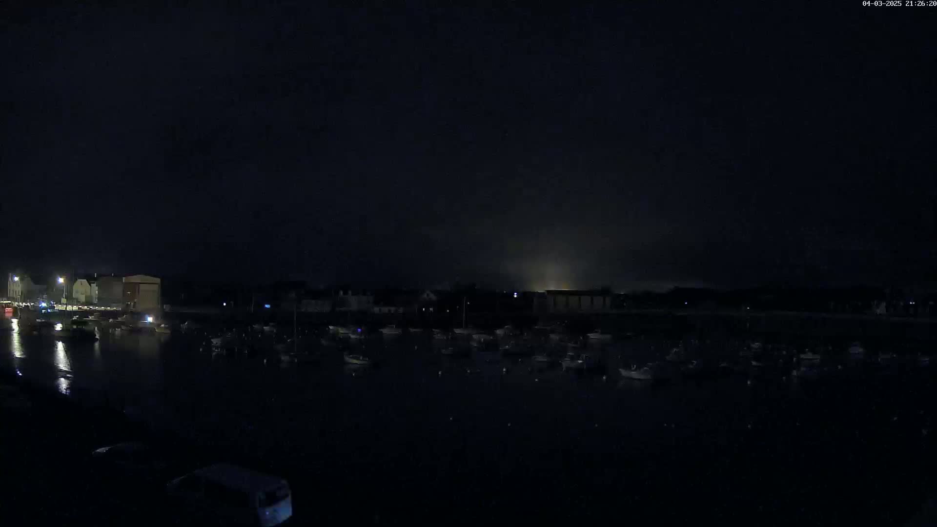 A dark nighttime harbor scene shows numerous boats moored in calm water, with dimly lit buildings visible along the shoreline under a mostly clear, dark sky.