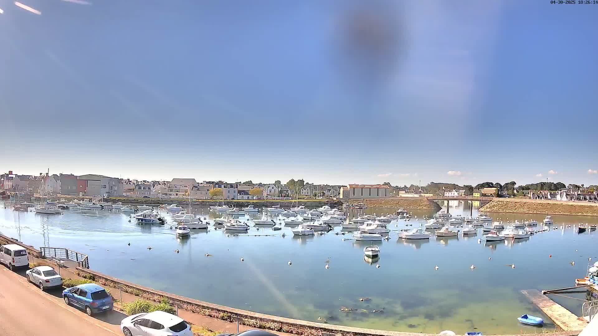 Under a clear blue sky, a calm harbor is filled with numerous docked boats, surrounded by buildings and a stone embankment.