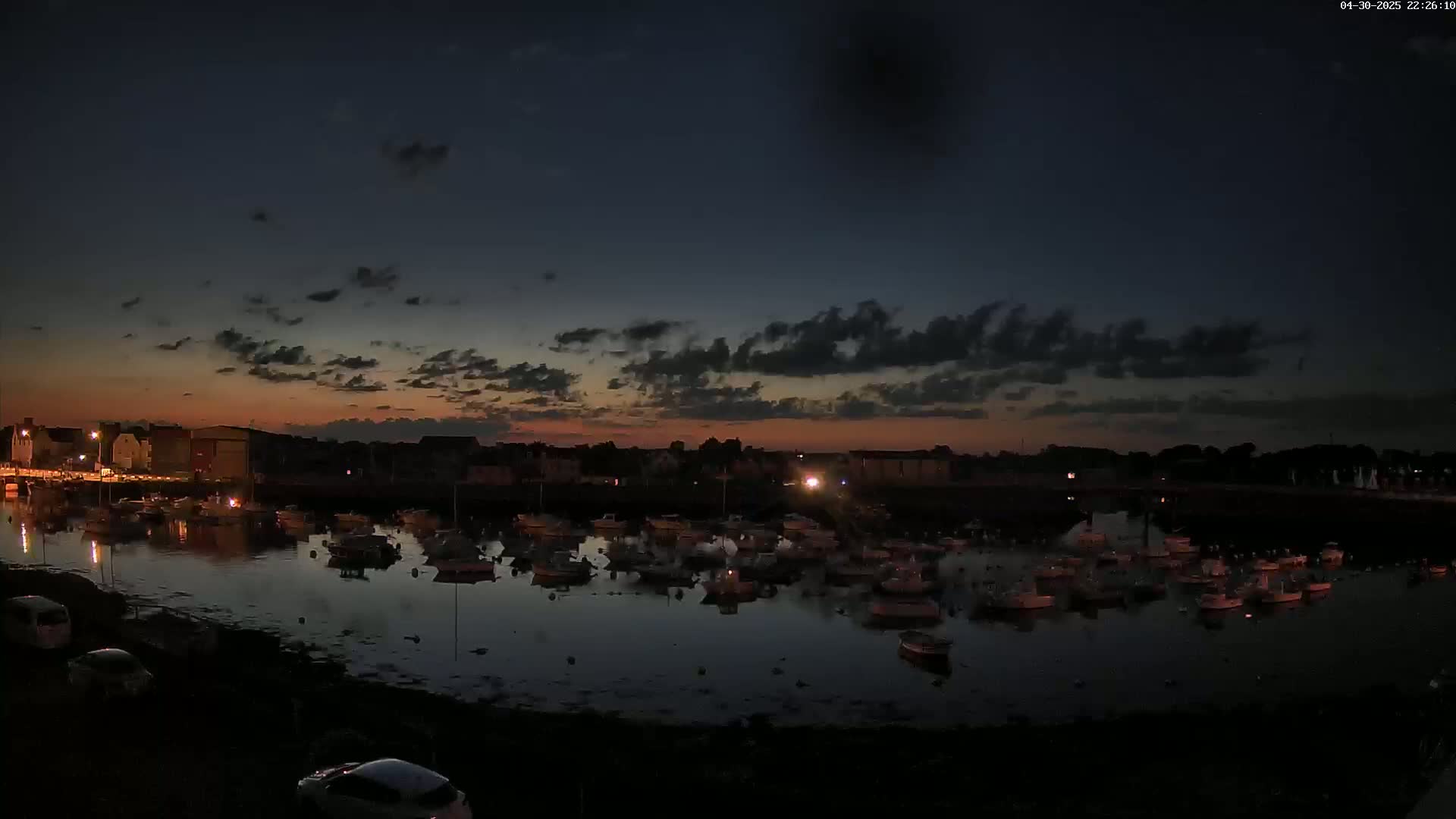 Many boats are moored in a calm harbor at twilight under a partly cloudy sky.