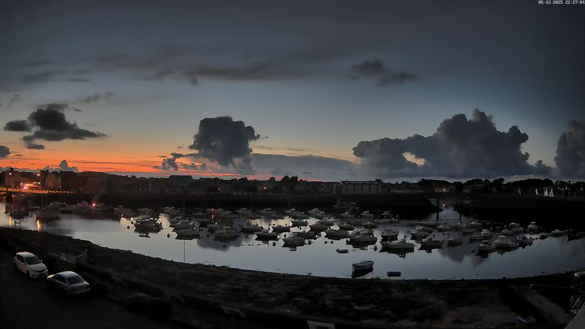 Many boats are moored in a calm harbor at twilight under a partly cloudy sky with streaks of orange and pink on the horizon.
