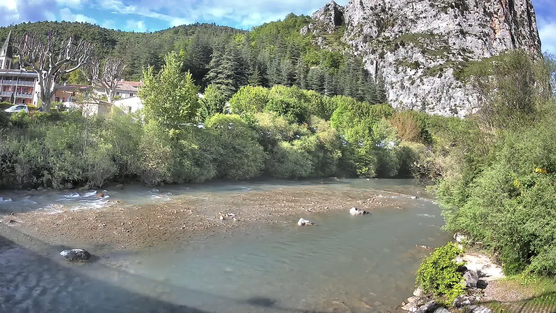 A partly cloudy day shows a shallow river flowing past a small village nestled at the base of a steep, rocky cliffside, surrounded by lush green vegetation.