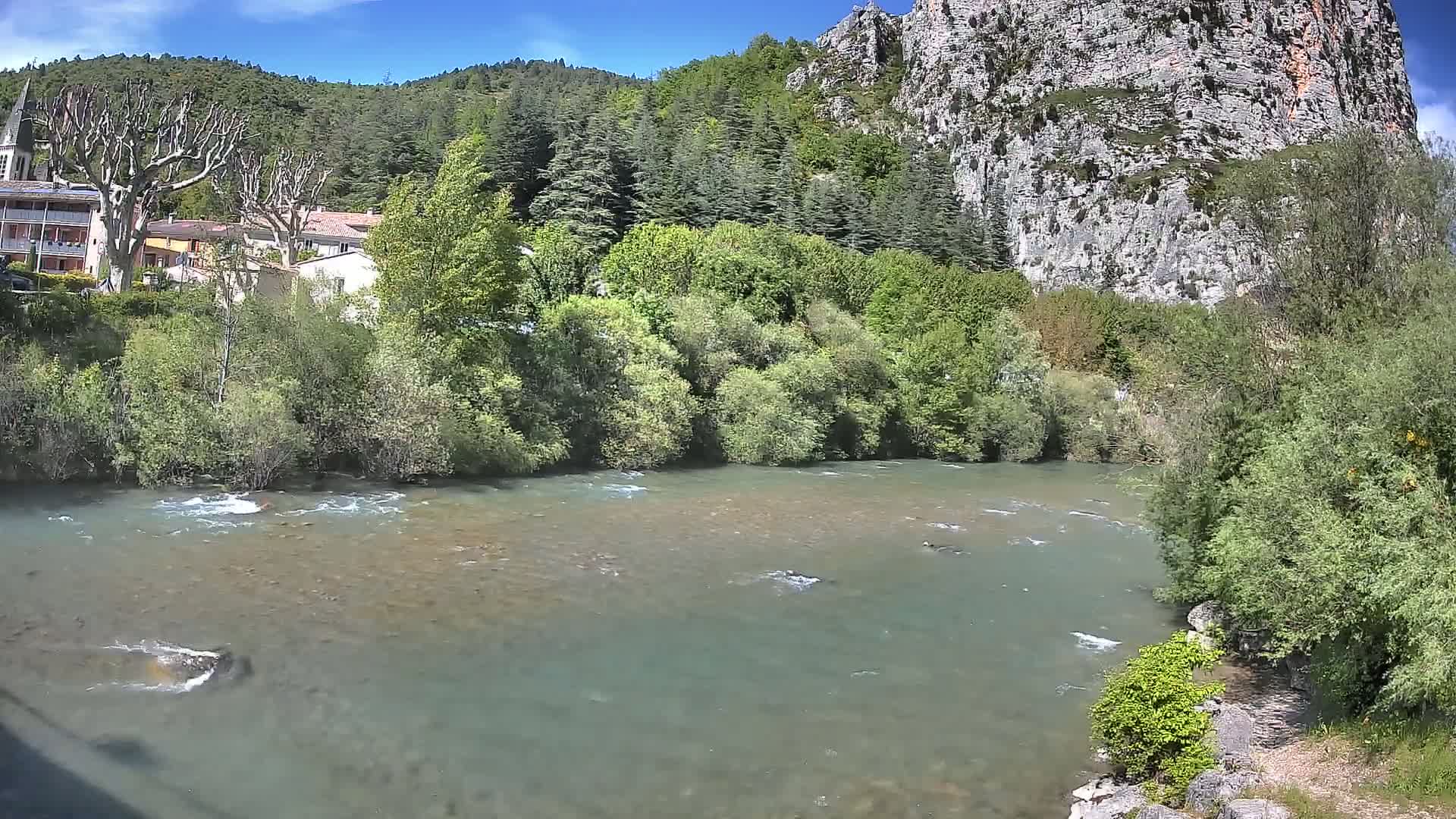 A clear, shallow river flows past a village nestled at the base of a rocky cliff, under a mostly sunny sky.