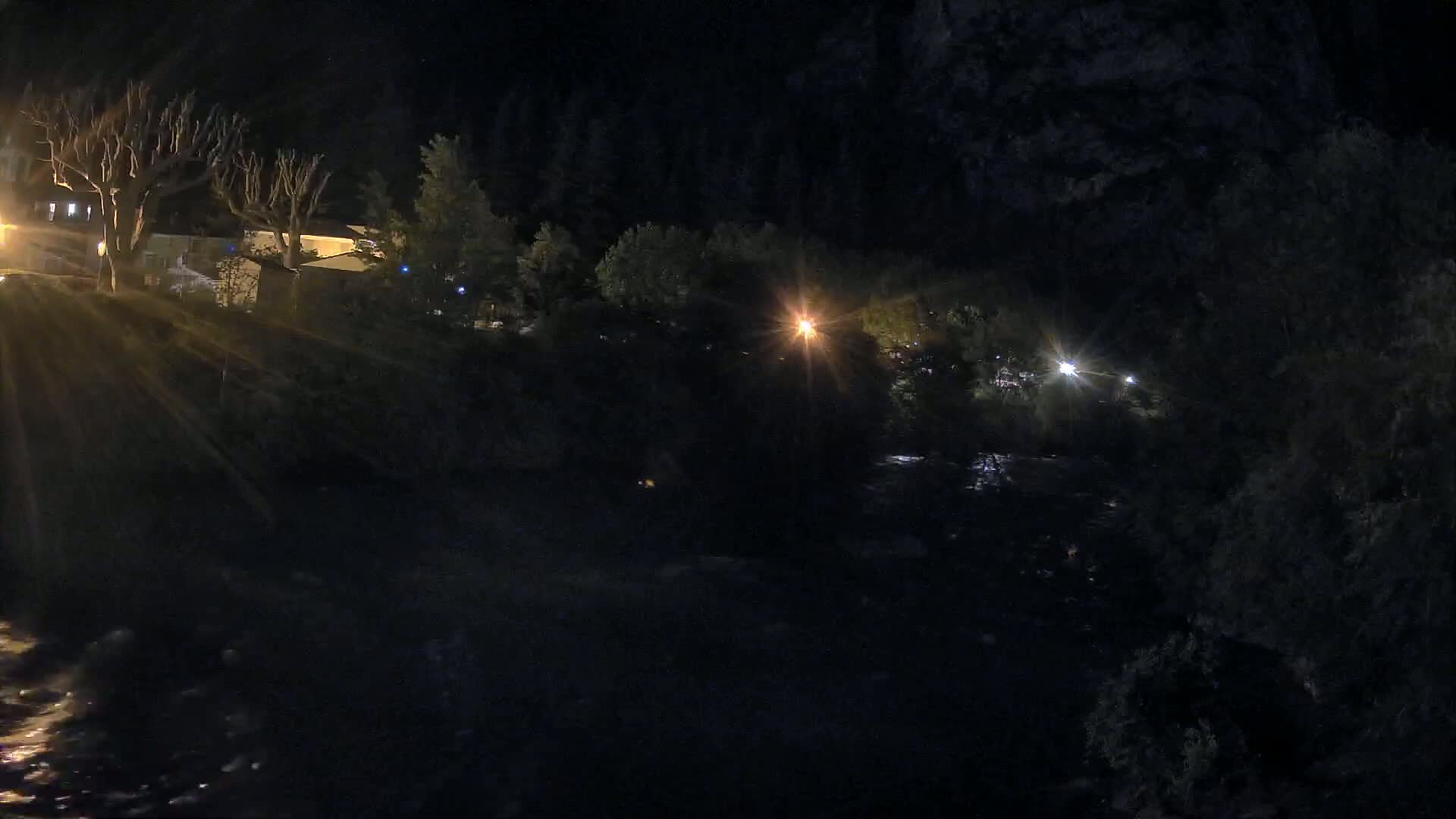 At night, a dark river flows beneath a hill with sparsely lit buildings and trees.
