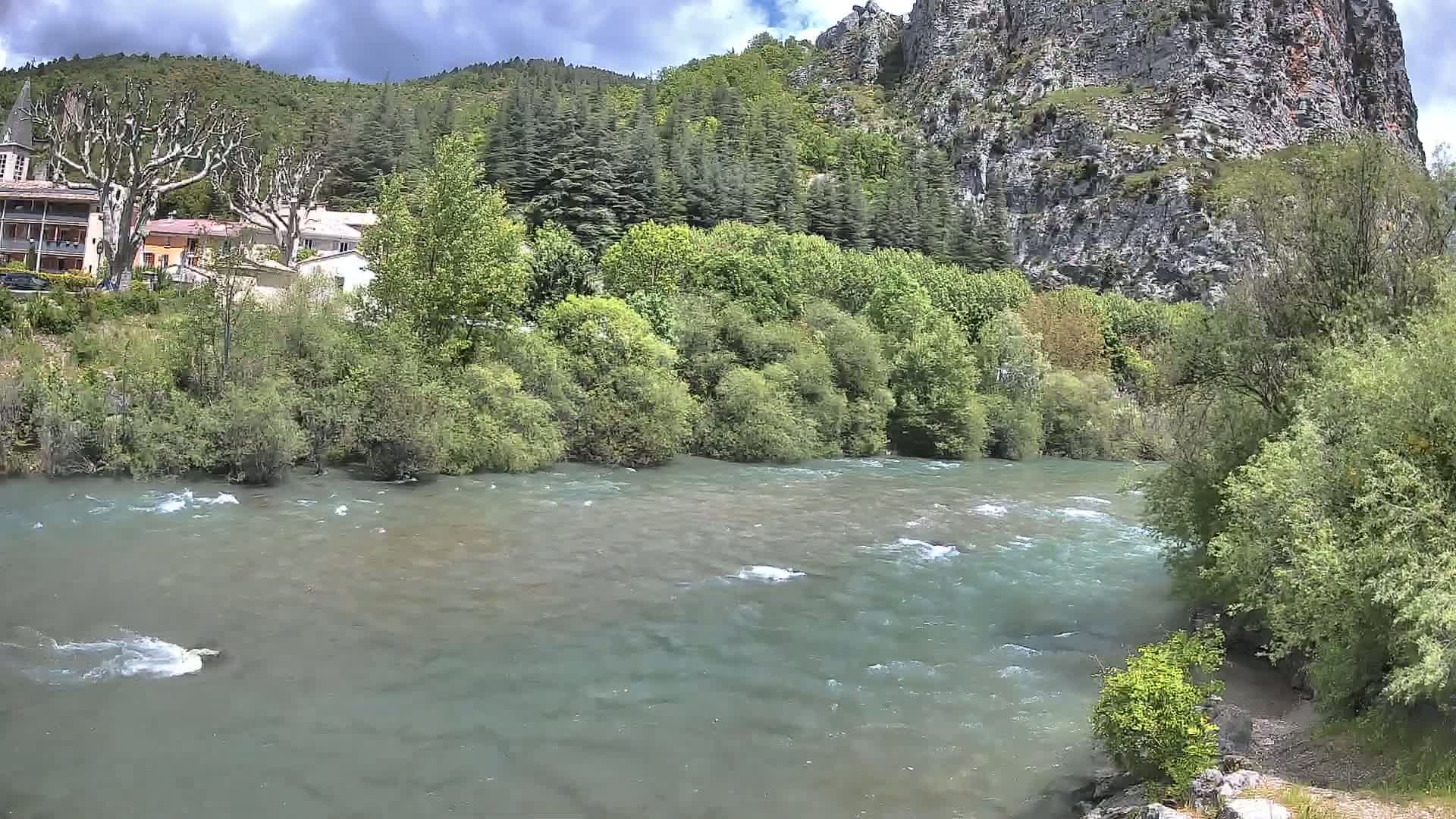 A swiftly flowing river under a partly cloudy sky, bordered by lush green vegetation and a rocky cliff face overlooking a small village.