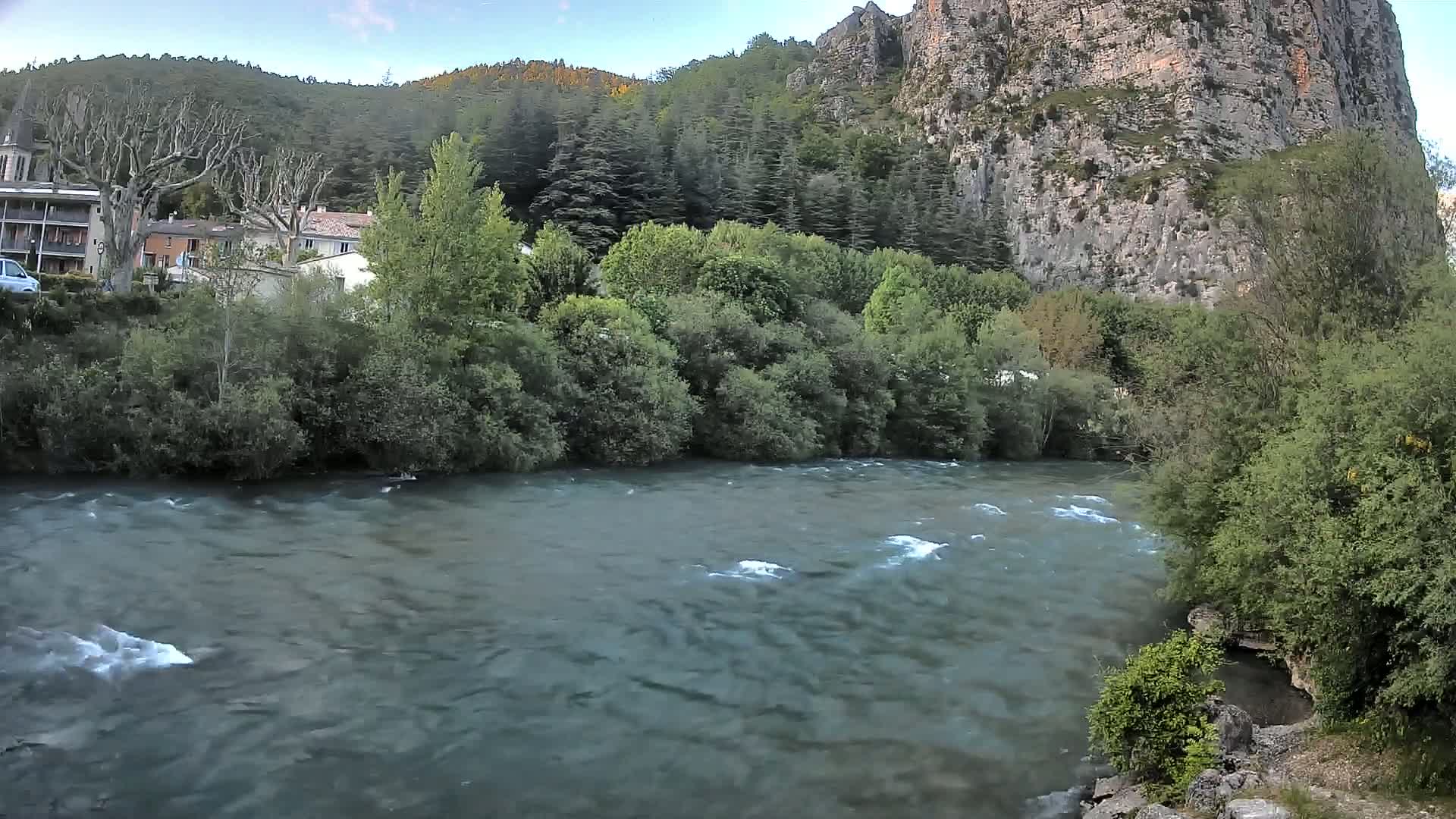 A swiftly flowing river runs past lush green vegetation and a village nestled beside a steep, rocky cliff under a partly cloudy sky.