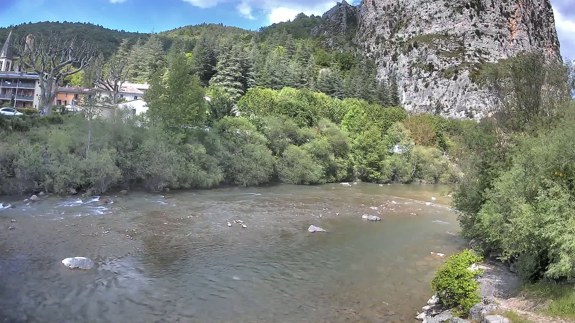 A moderately flowing river runs through a lush green valley, alongside a small village nestled against a steep, rocky cliff face under a partly cloudy sky.