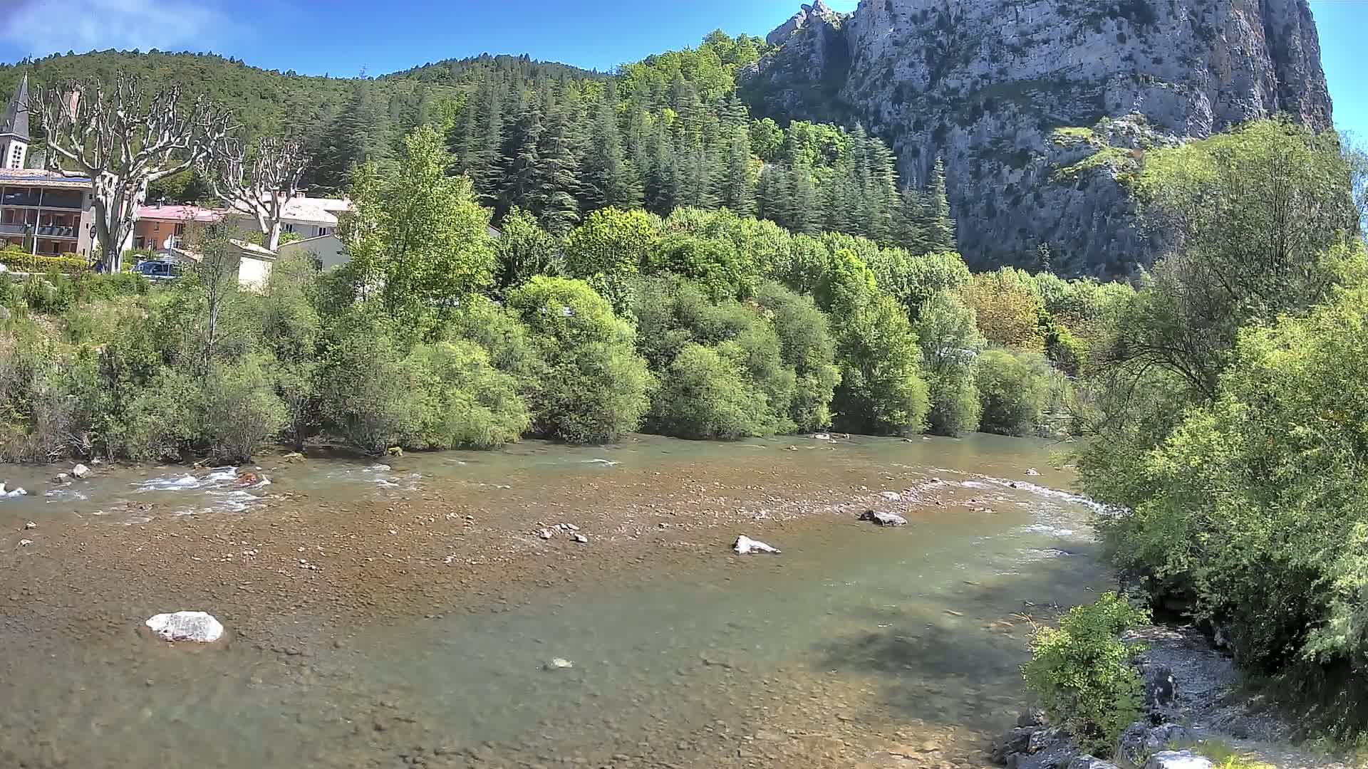 A shallow, rocky river flows past lush green vegetation and a small village nestled against a steep, rocky hillside under a sunny sky.