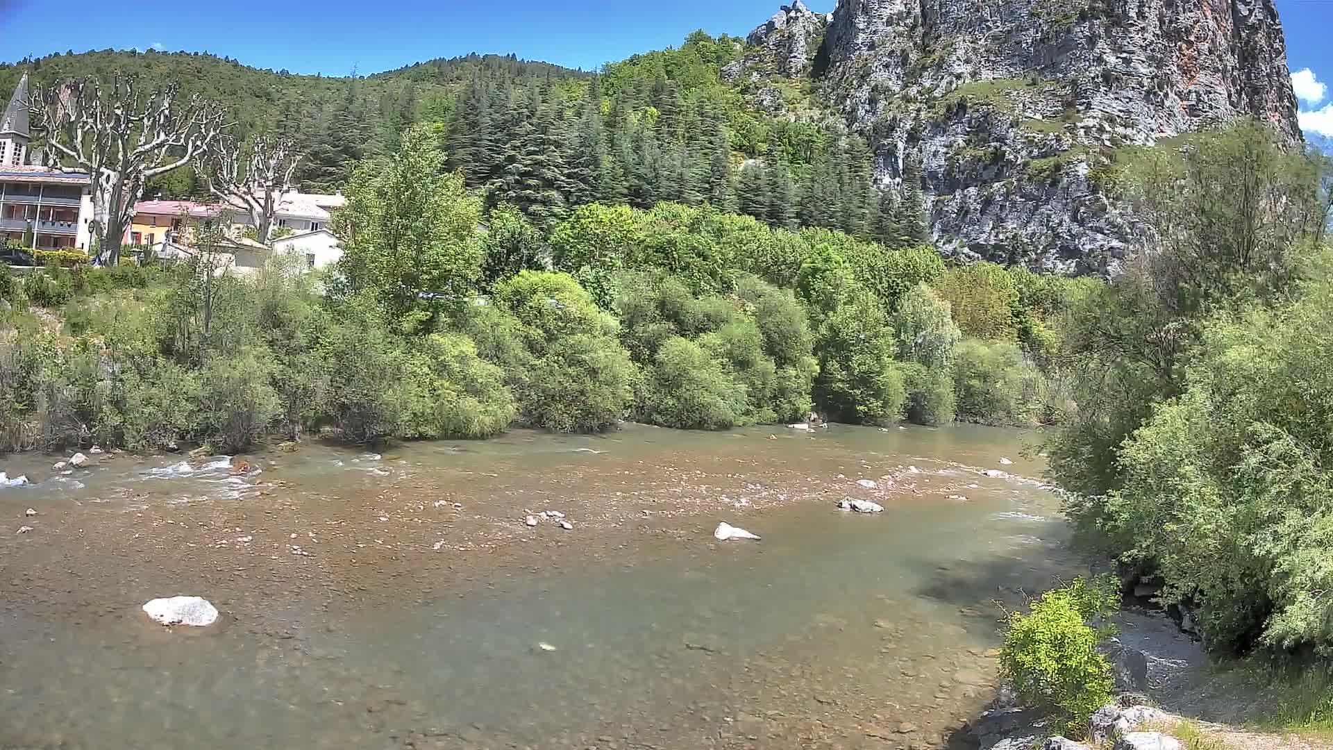 A clear river flows past lush green vegetation and a village nestled against a rocky cliff face under a sunny sky.