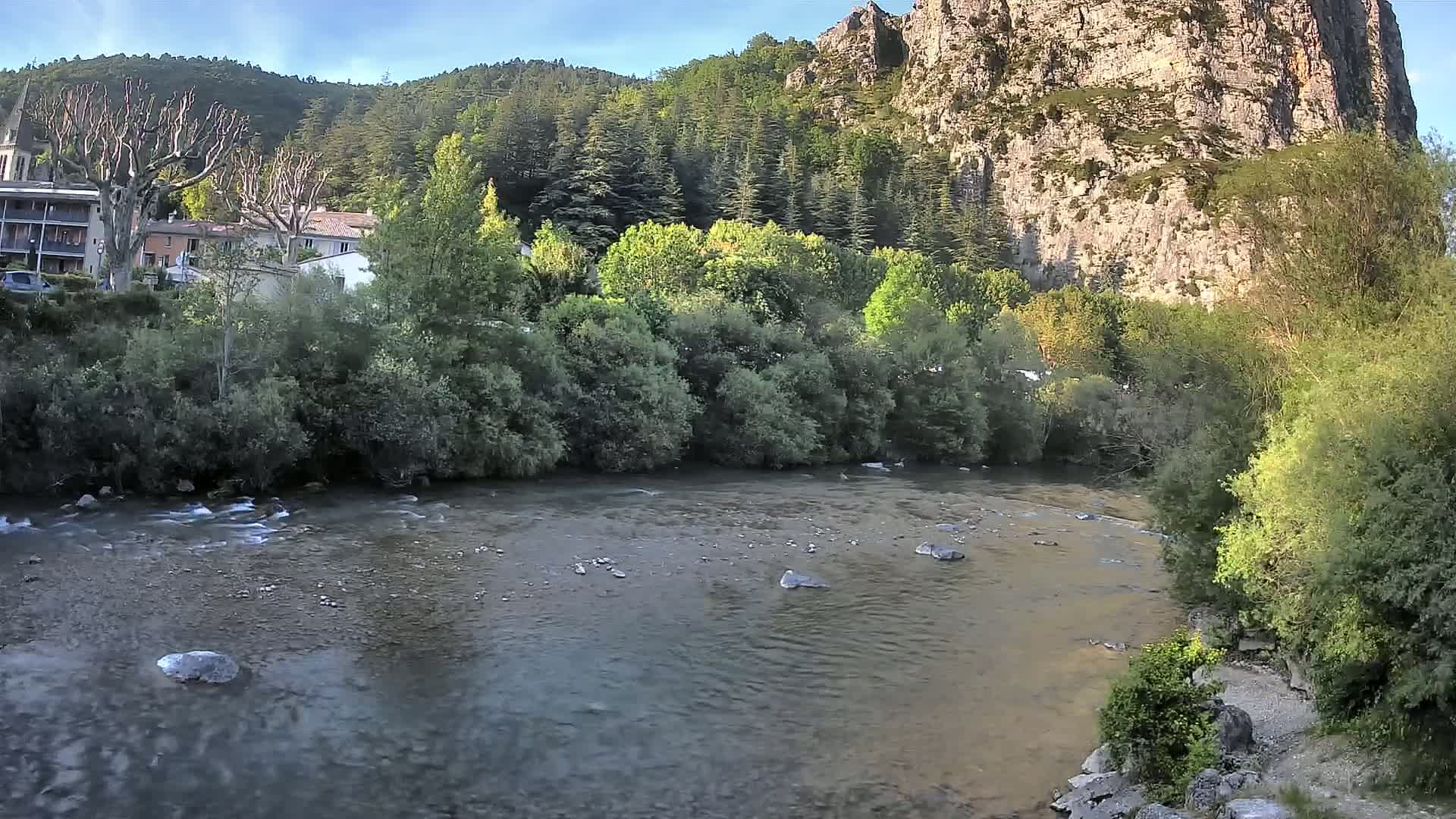A calm river flows past lush green vegetation and buildings nestled at the base of a large, rocky cliff under a clear sky.