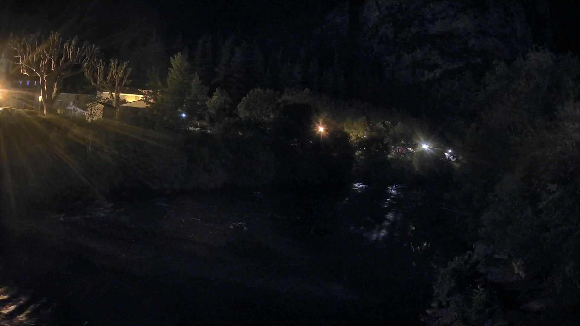 At night, a dark river flows past buildings and trees illuminated by scattered lights.