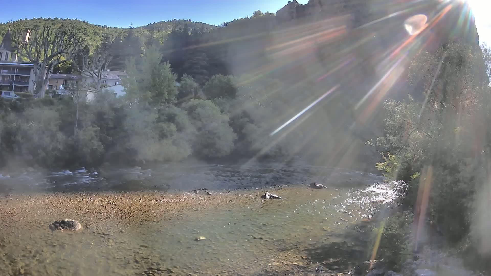 A sunlit shallow river flows past a small town nestled in a valley, surrounded by lush green hills under a clear blue sky.