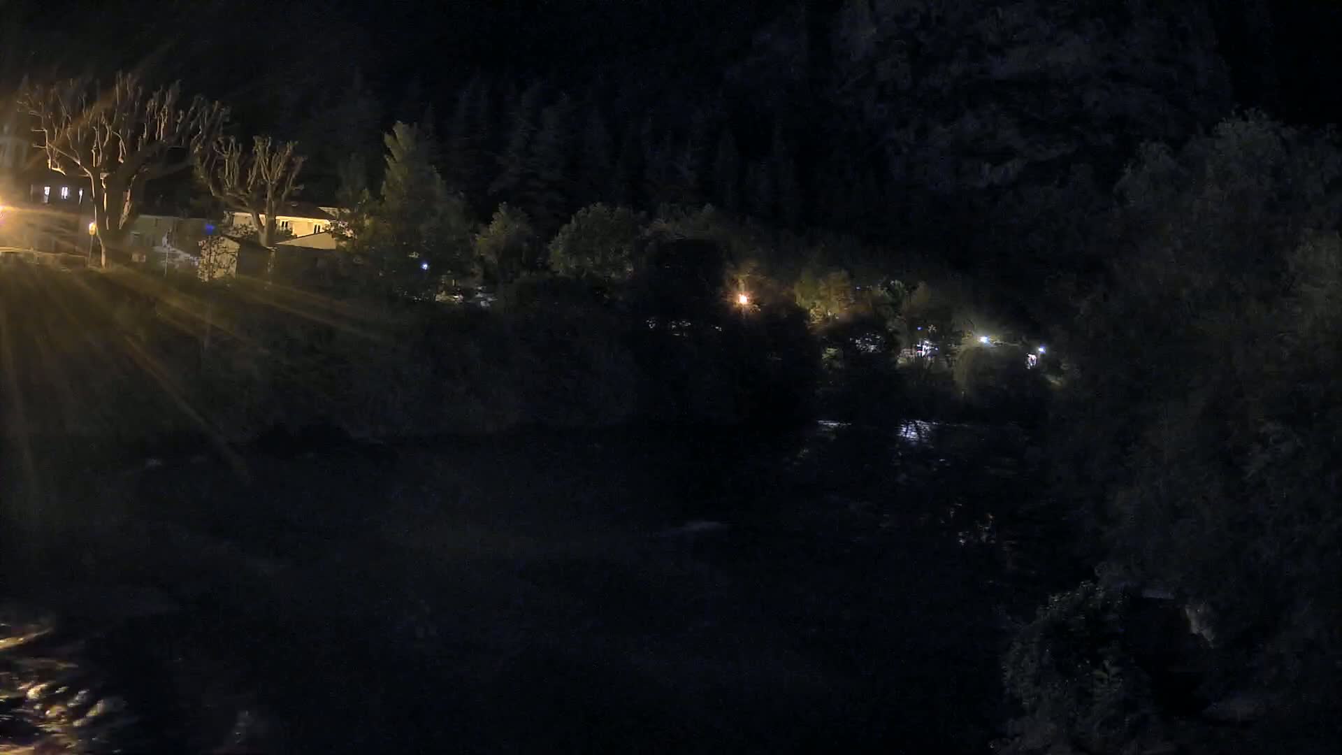 A nighttime view of a dark river flowing past a line of houses and trees, illuminated by scattered lights.