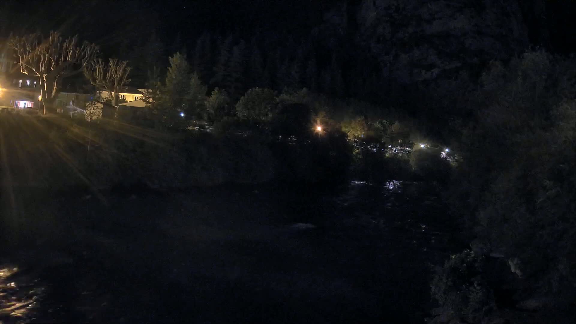 At night, a dark river flows past sparsely lit buildings and trees under a clear, dark sky.