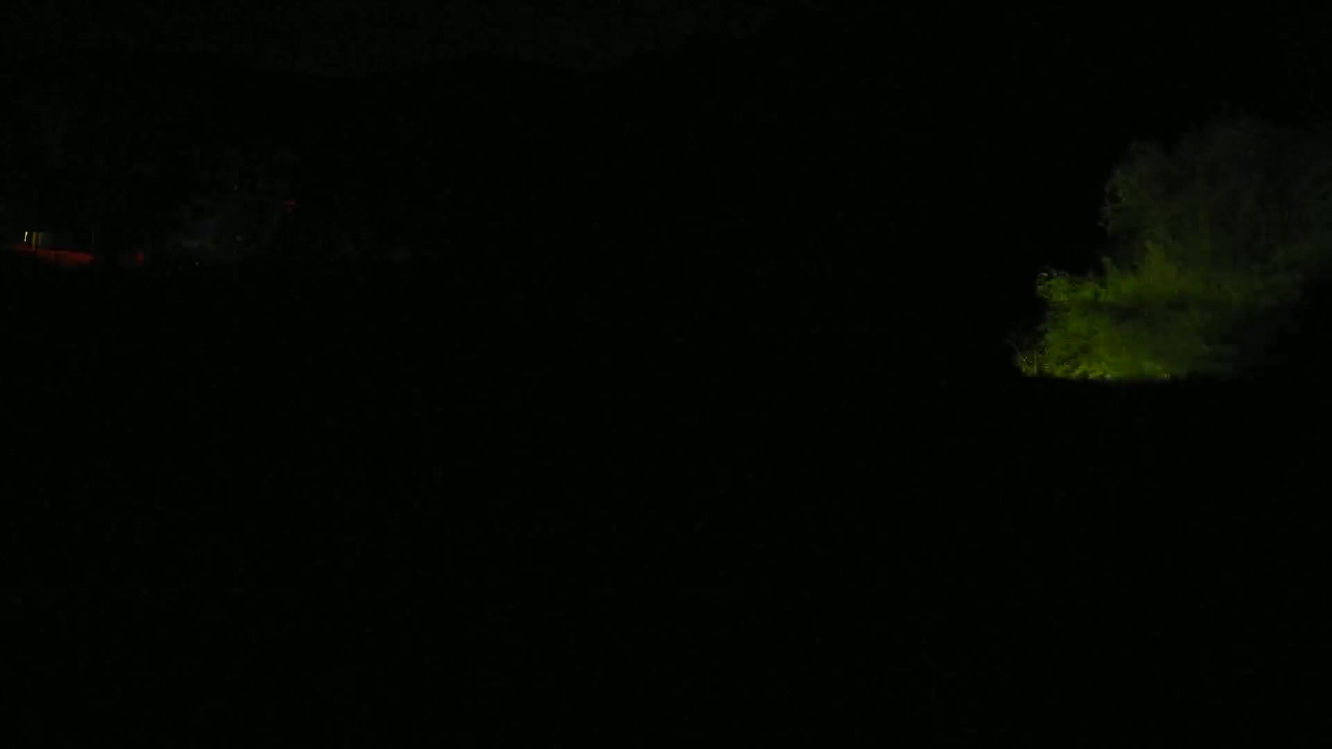 A dimly lit, nighttime scene shows a vaguely shaped, glowing green object on the right and faint red lights on the left.