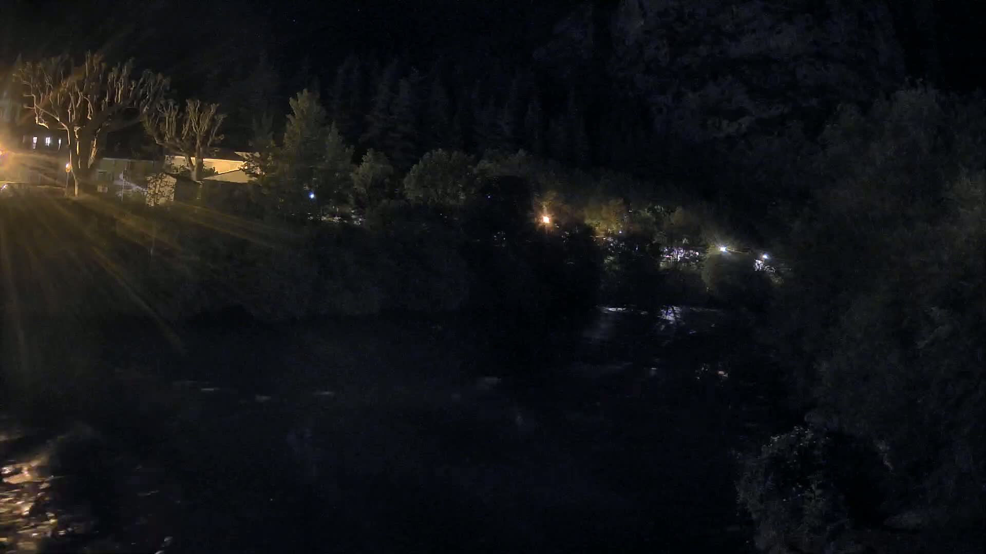 A dark river flows under a nighttime sky past trees and buildings with scattered lights.