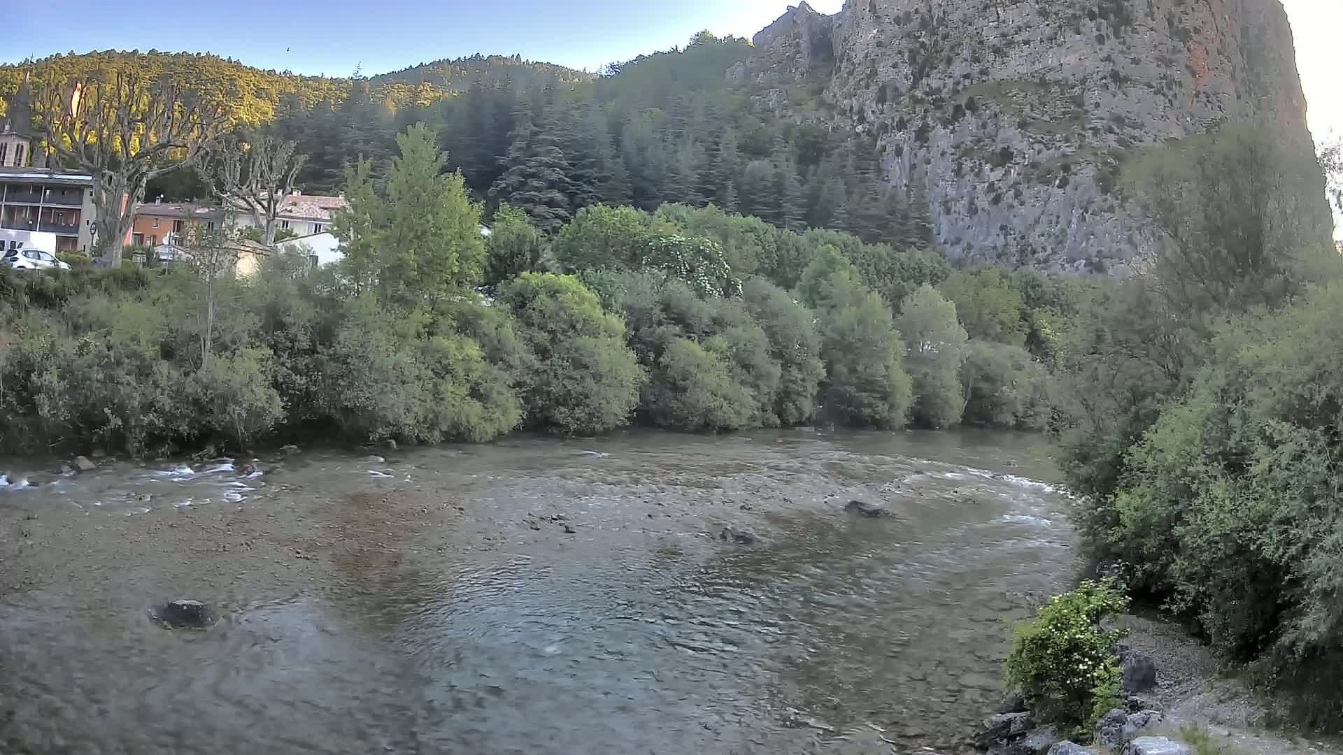 A calm river flows past lush green vegetation and a village nestled at the base of a large, rocky cliff under a clear sky.