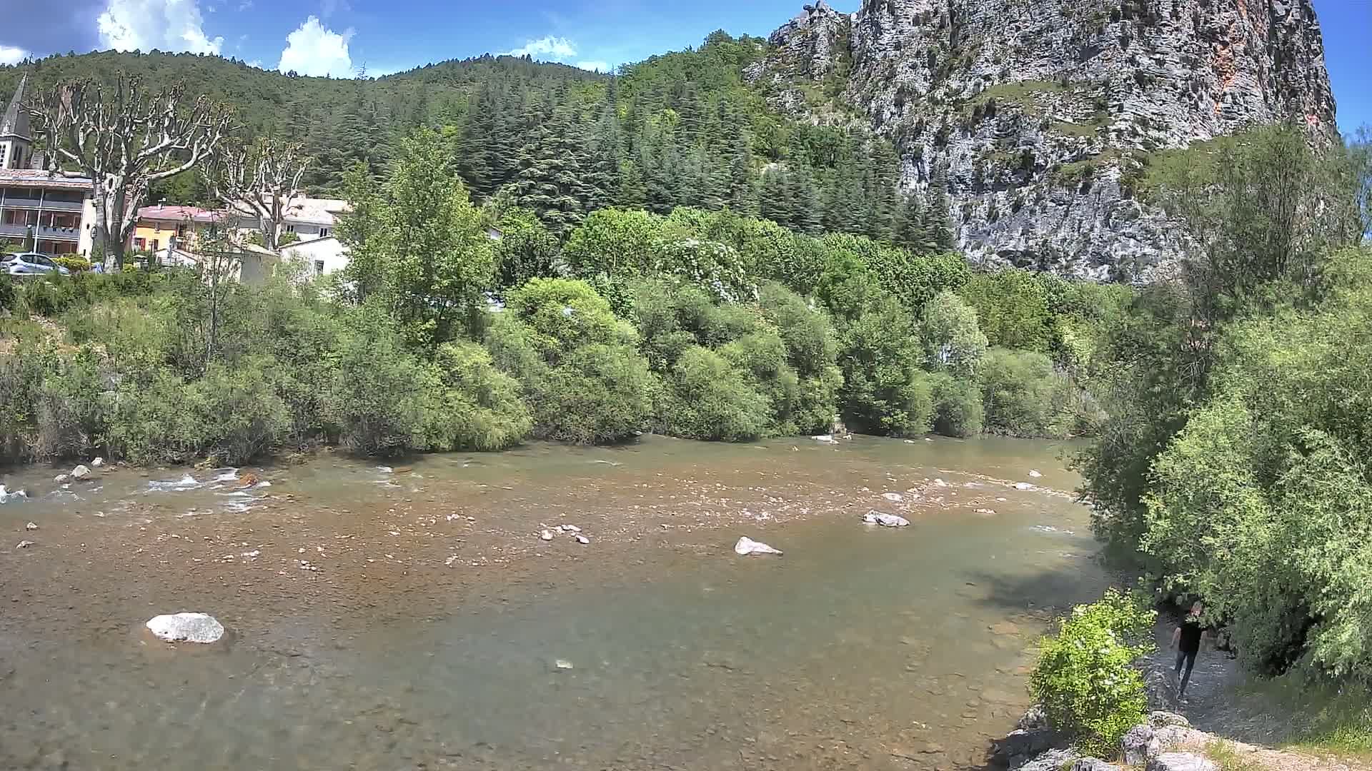 A shallow, rocky river flows through a lush green valley under a partly cloudy sky, with a village and a large rock face visible in the background.