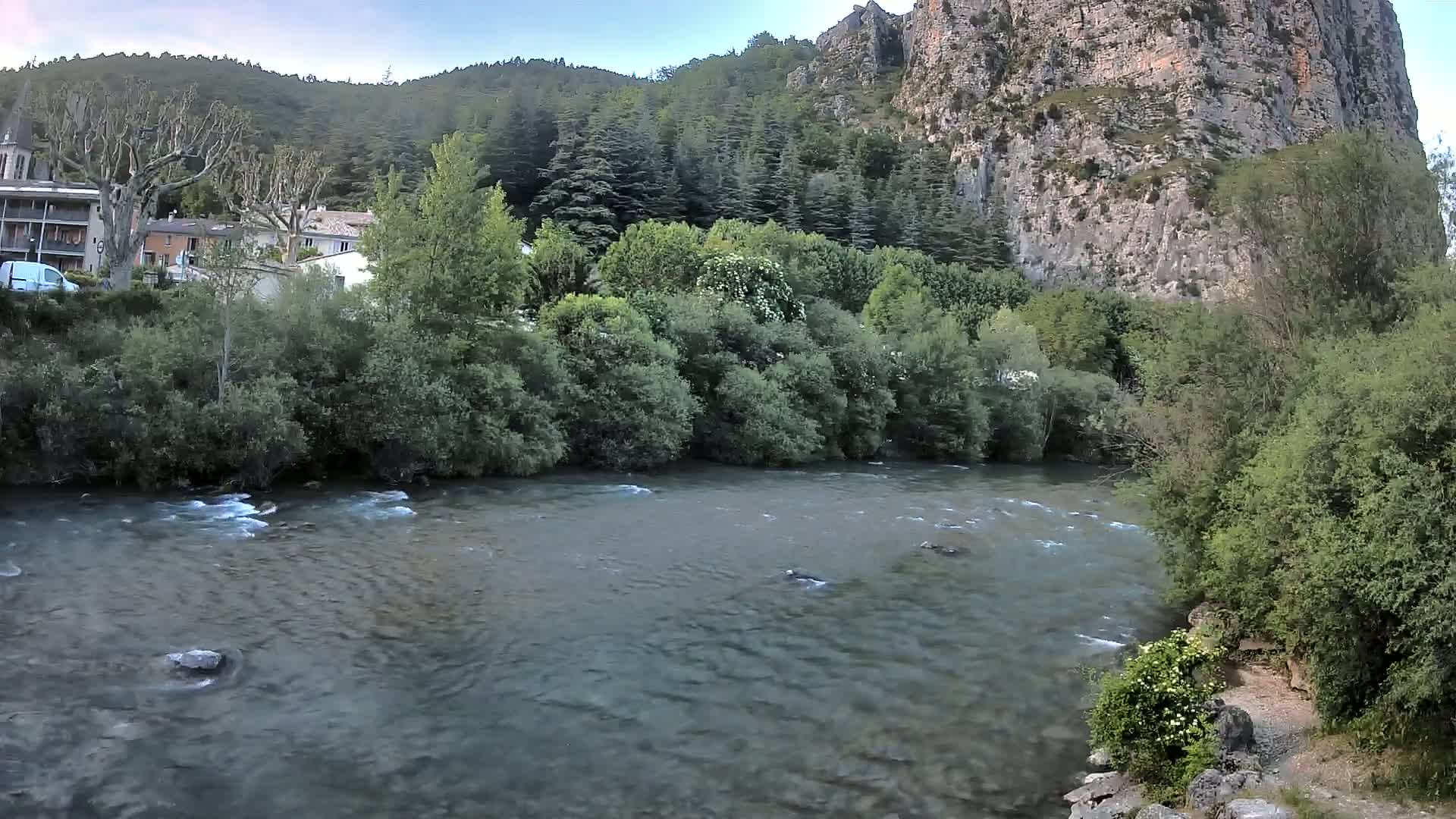 A river flows gently past lush green vegetation and buildings nestled at the base of a large, rocky cliff under a partly cloudy sky.