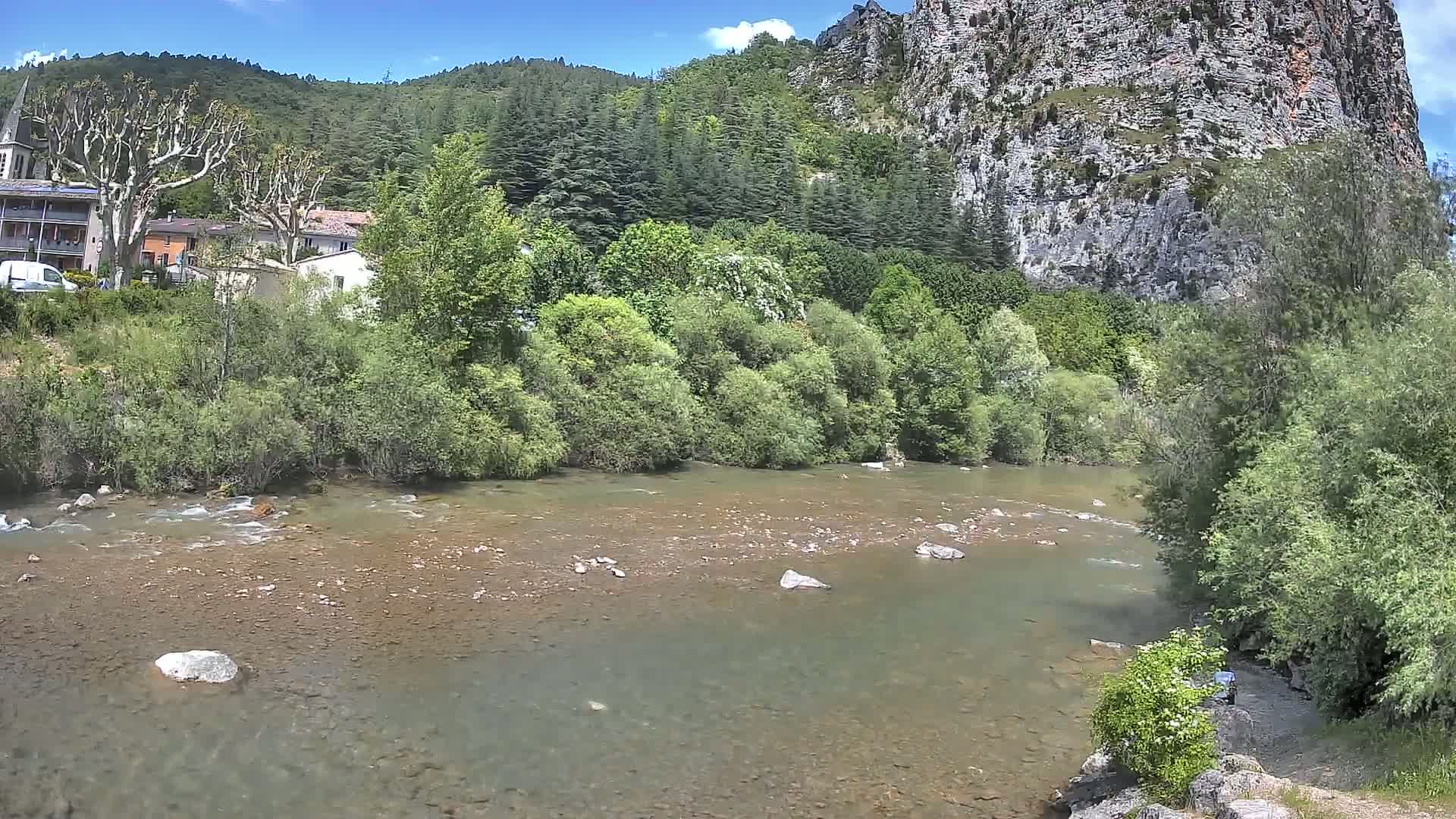 A shallow, rocky river flows past a village nestled at the base of a steep, rocky cliff, under a sunny sky.