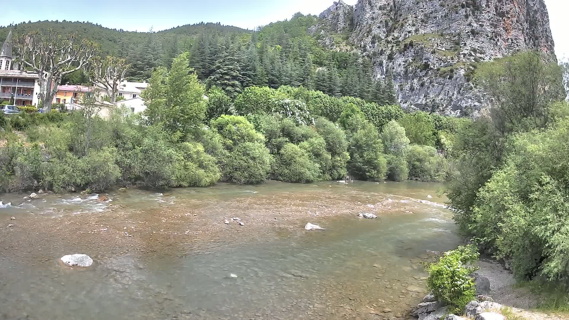 A shallow, rocky river flows past lush green vegetation and a village nestled beneath a steep, rocky cliff face on a partly cloudy day.