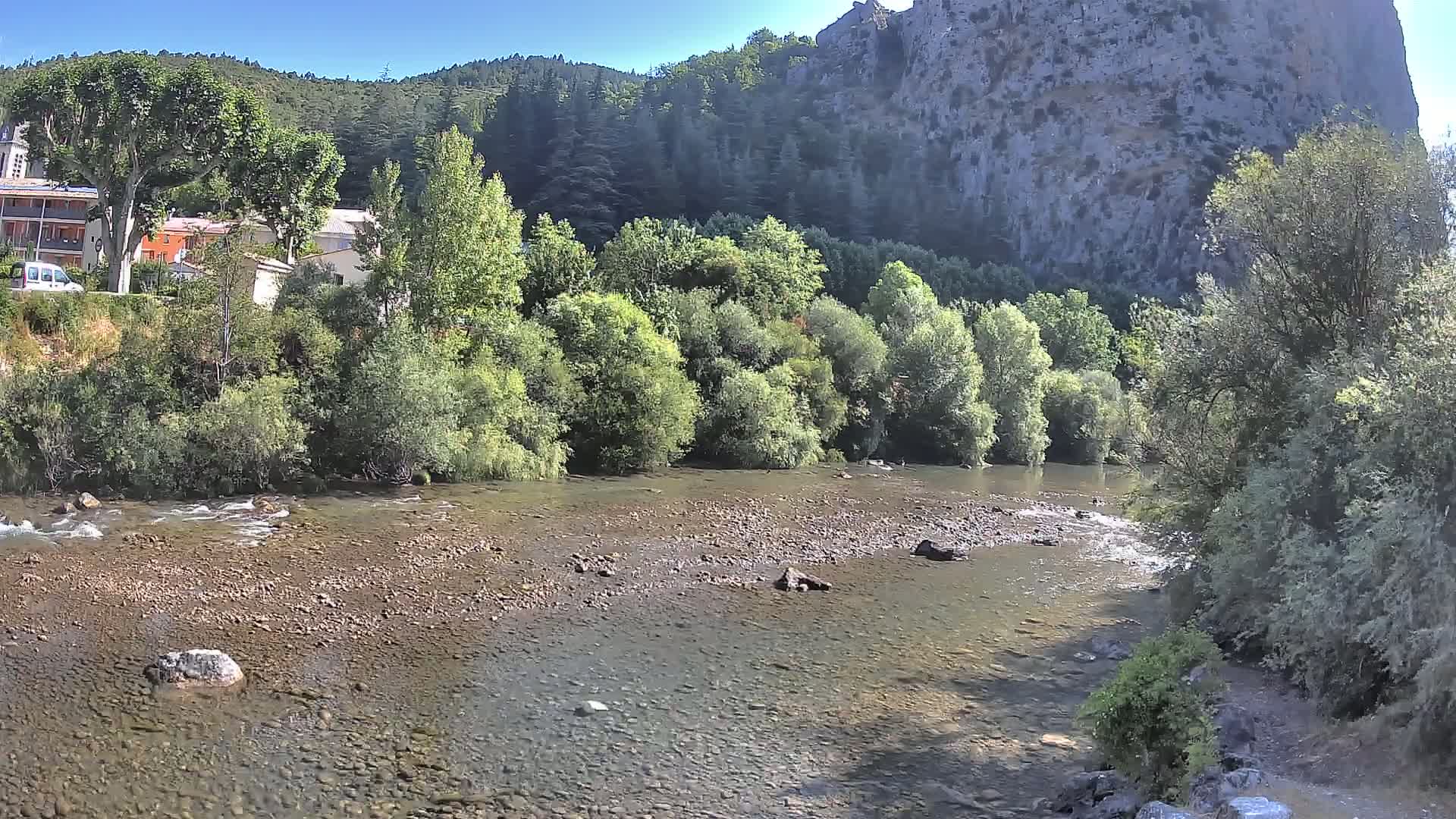 A shallow, rocky river flows past lush green vegetation and a village nestled beneath a large, light-grey cliff face on a sunny day.