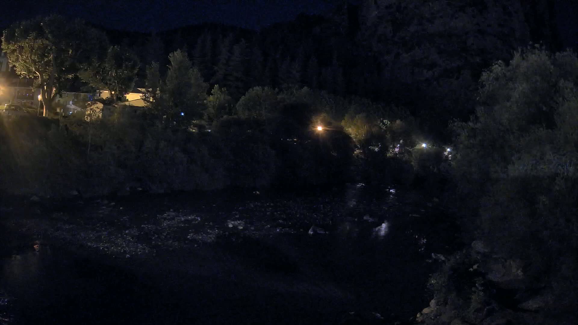 A dark river flows under a nighttime sky past a tree-lined bank with buildings and lights visible in the distance.