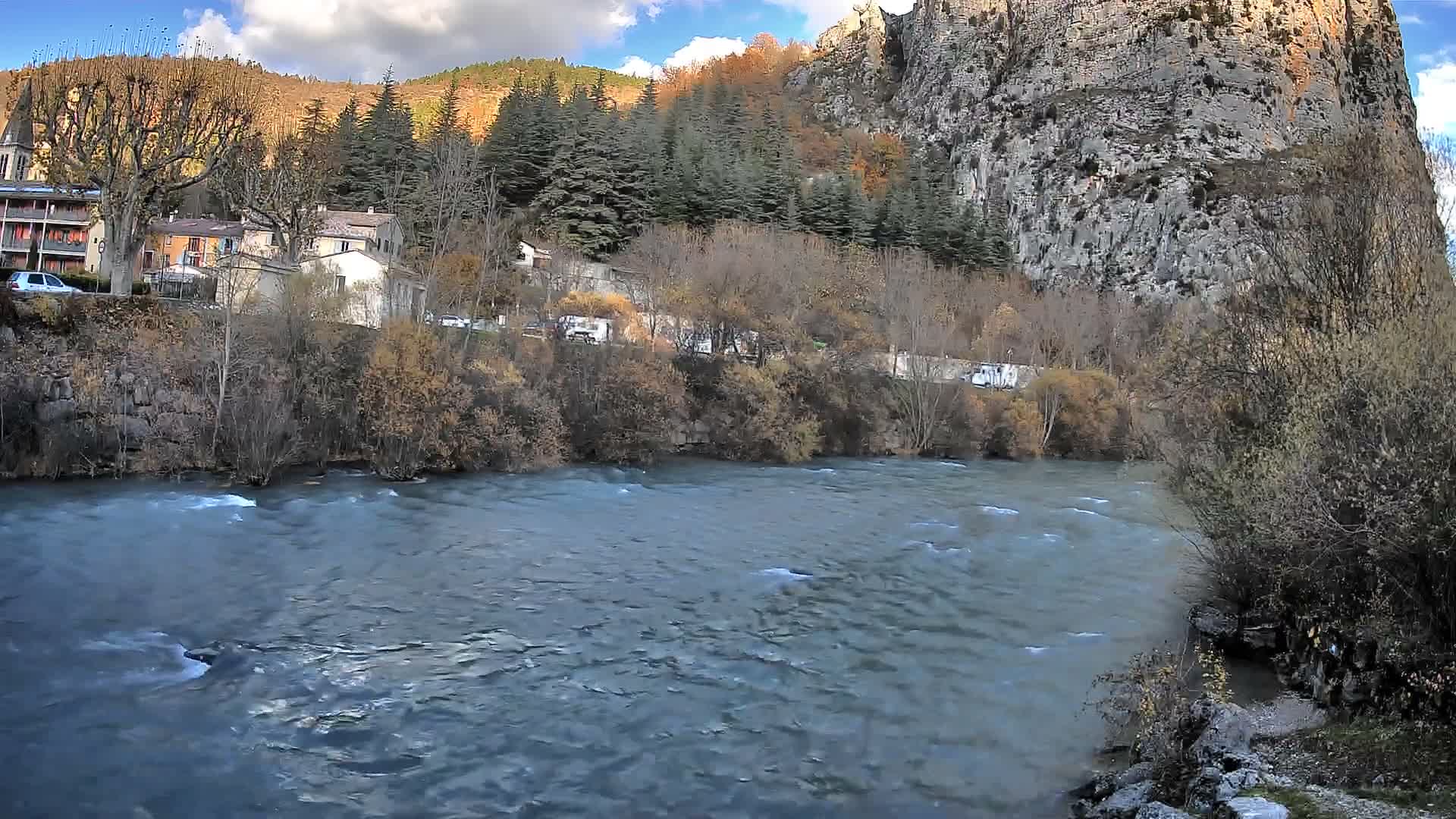 A dark river flows under a nighttime sky past a tree-lined bank with buildings and lights visible in the distance.