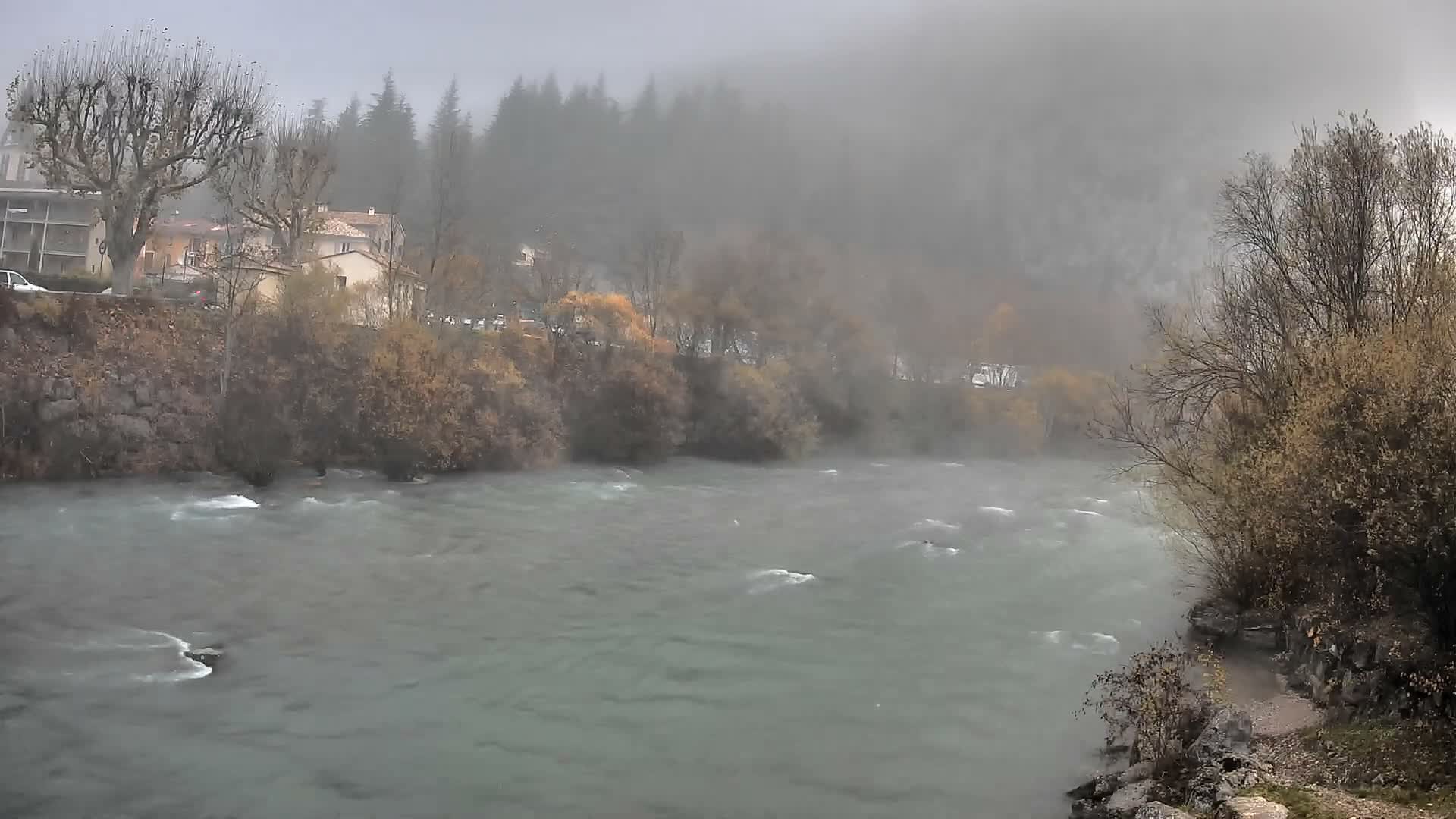 A wide, turbulent river flows through a misty valley under an overcast sky, flanked by bare trees and autumn-toned shrubs, with buildings visible on the left bank and forested hills fading into the heavy fog in the background.