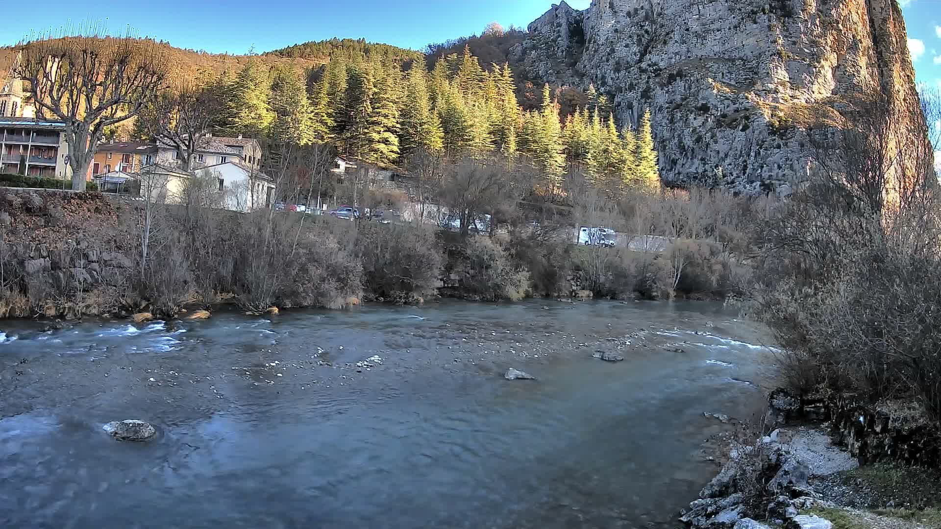 A wide river with some rapids flows through a valley featuring buildings and large bare trees on the left bank, a road with vehicles, a dense coniferous forest, and a towering rocky cliff on the right, all under a clear, sunny sky.
