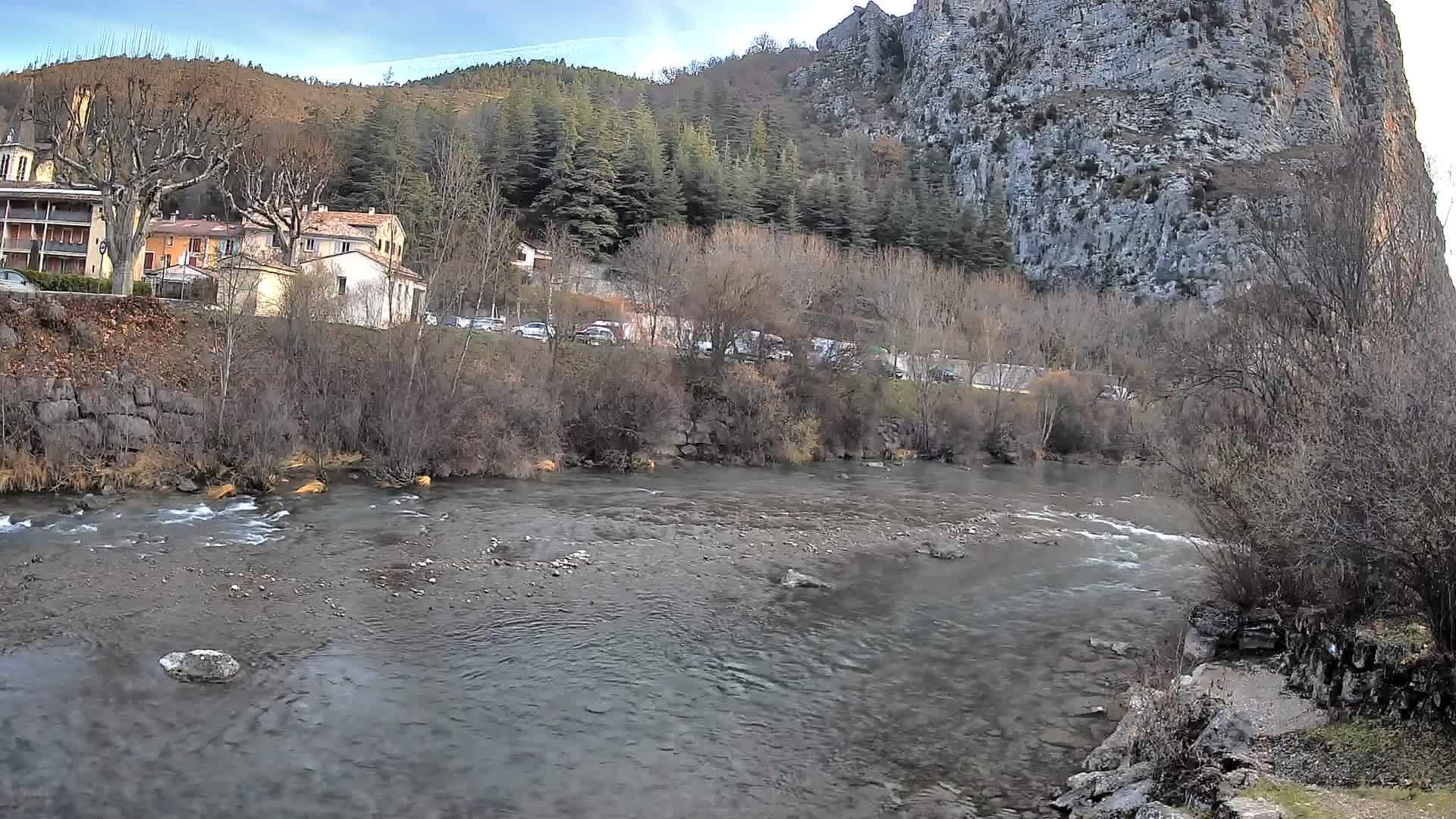 A wide river flows through a valley, flanked by buildings and bare trees on the left bank and a towering rocky cliff on the right, all under a clear blue sky with a hint of cloud.