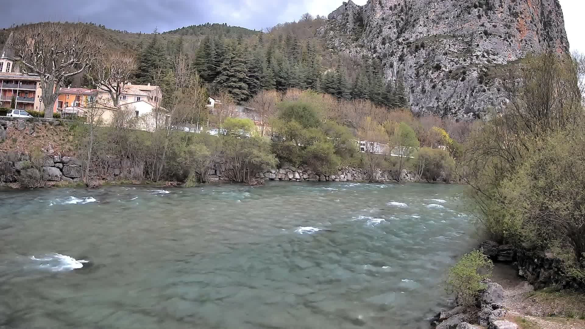 A wide river flows through a valley, flanked by buildings and bare trees on the left bank and a towering rocky cliff on the right, all under a clear blue sky with a hint of cloud.