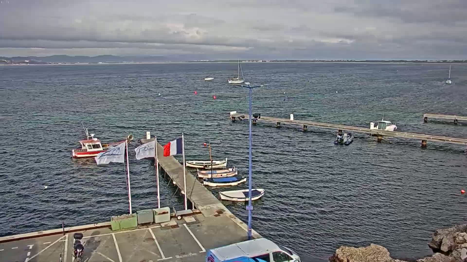 Several small boats are moored at a pier in calm water under a cloudy sky, with a larger boat further out in the bay.