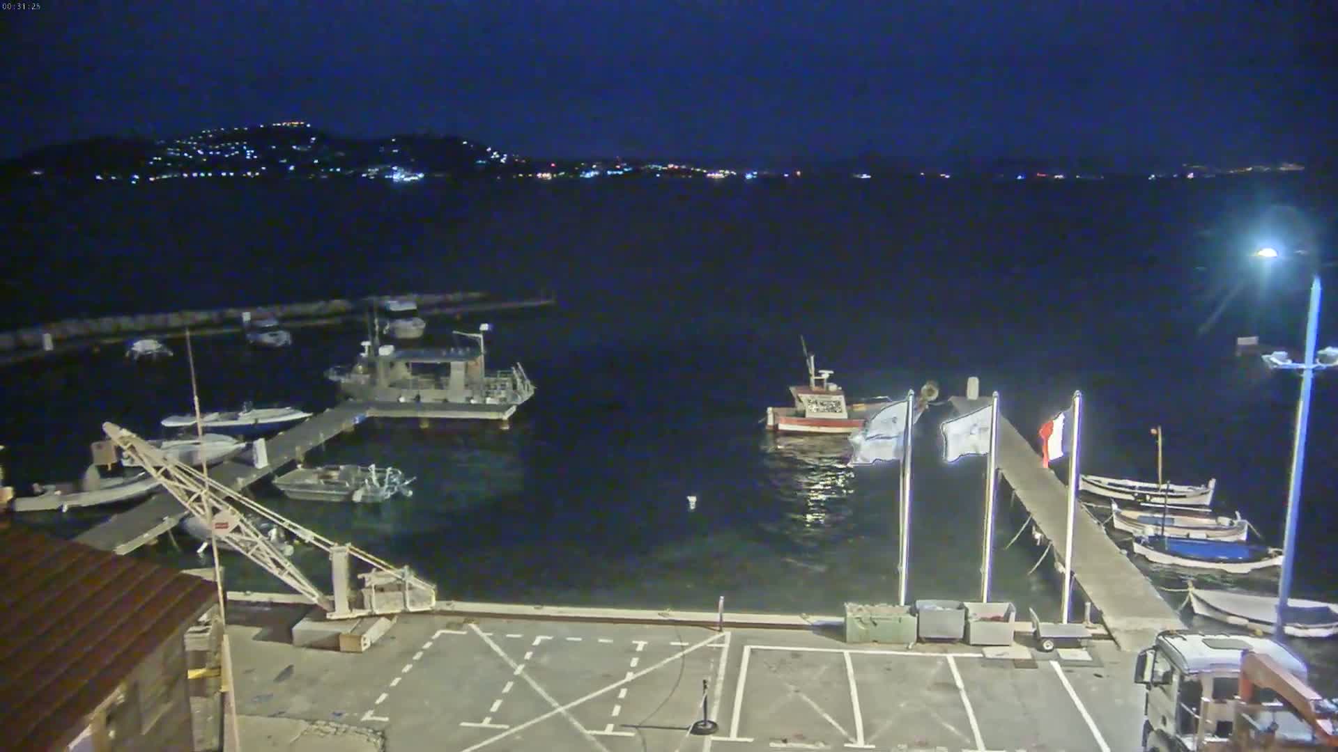 A nighttime view of a harbor with several boats moored at docks, a small crane, and a truck parked nearby, under a clear, dark sky.