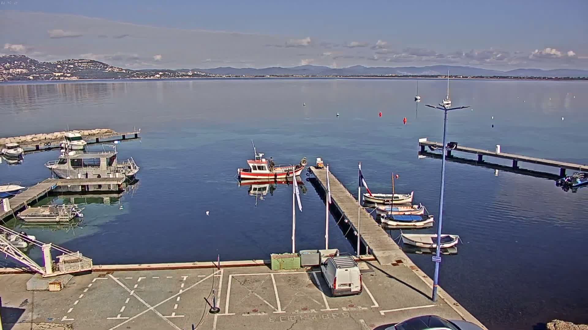 A calm, sunny day reveals a harbor with several boats moored at docks, a van parked nearby, and a distant mountain range visible across the water.