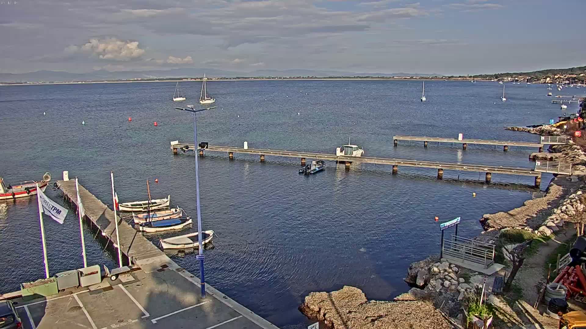 Several boats are moored at a small harbor with wooden piers extending into a calm body of water under a partly cloudy sky, with distant hills visible on the horizon.