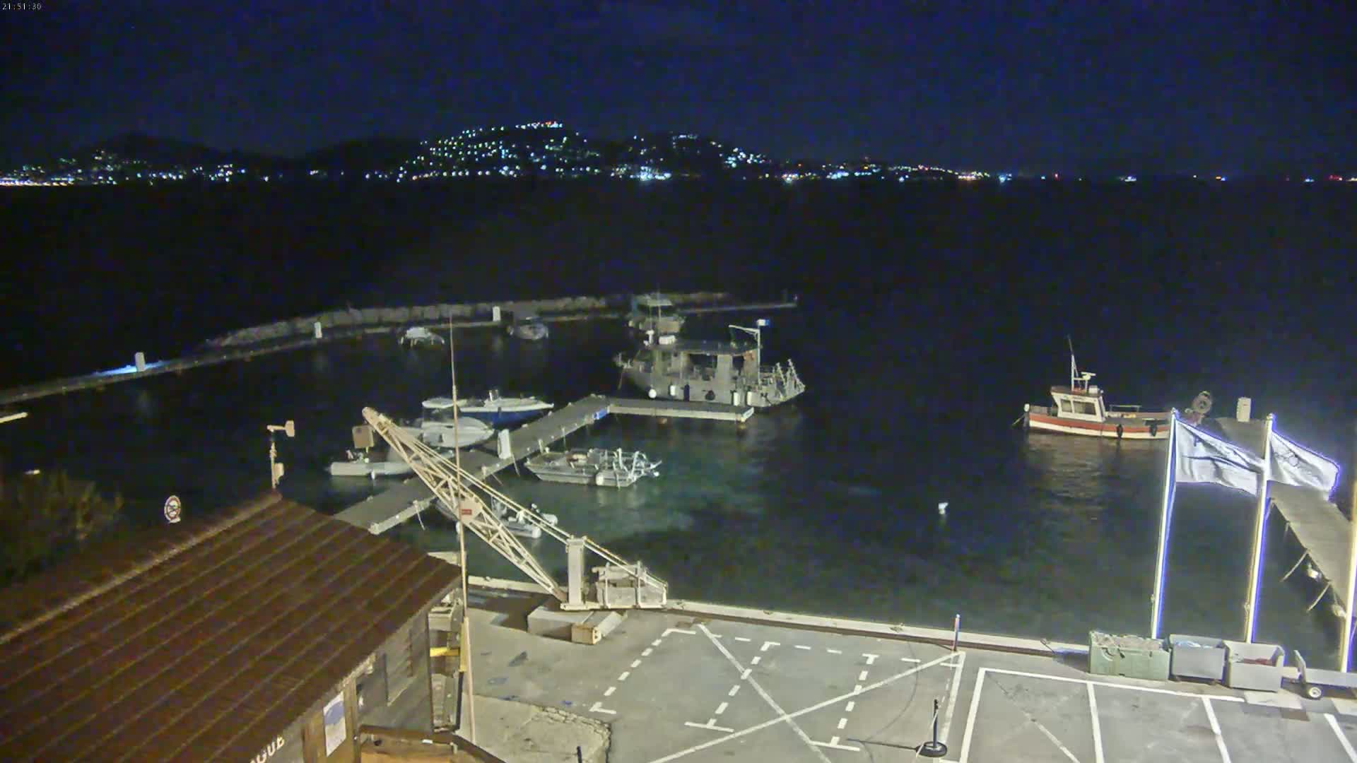 A nighttime view of a harbor with several boats docked, a crane, and a distant cityscape illuminated across the water under a clear, dark sky.