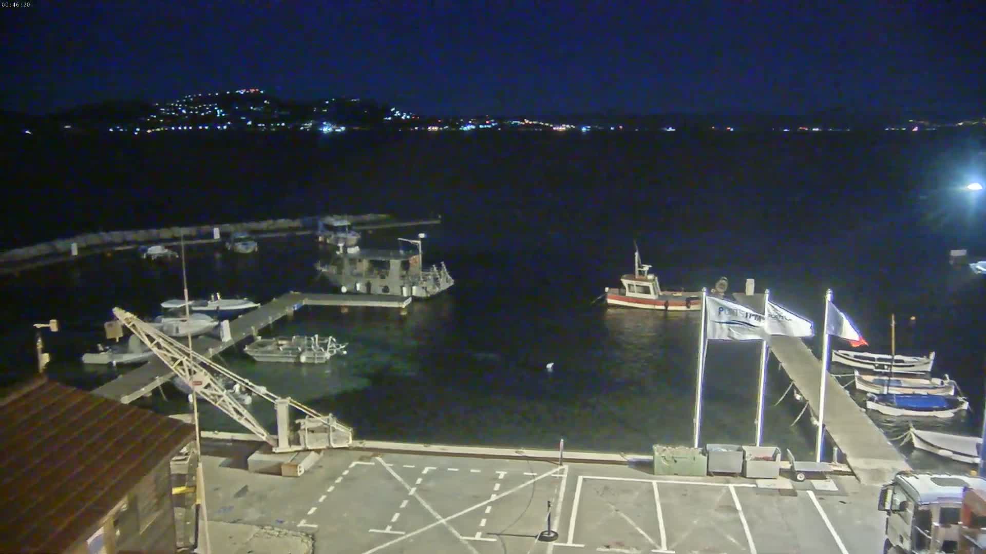 A nighttime view of a marina with several boats docked, a crane, and a distant city skyline illuminated across the water.
