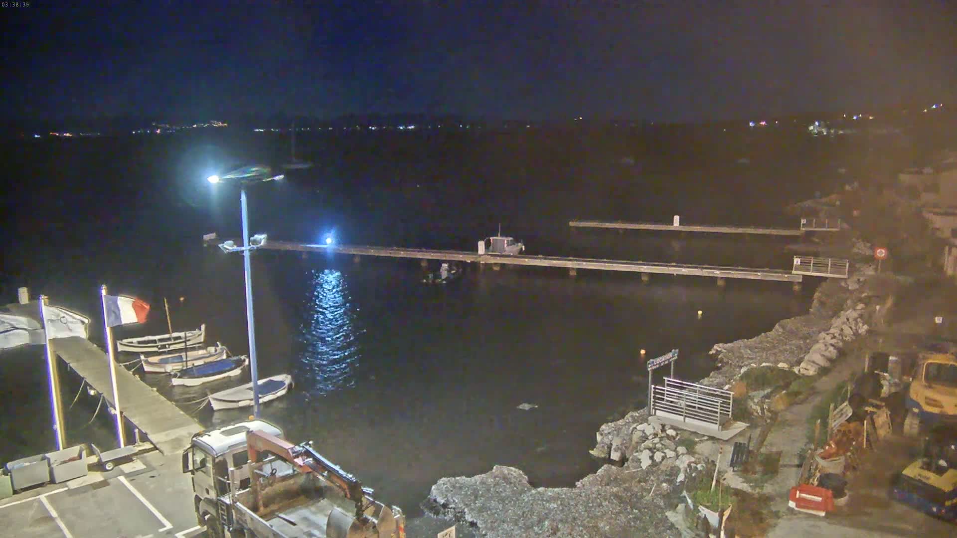 A nighttime view of a harbor with several small boats docked at piers, a truck, and construction equipment near a rocky shoreline under a dark, clear sky.
