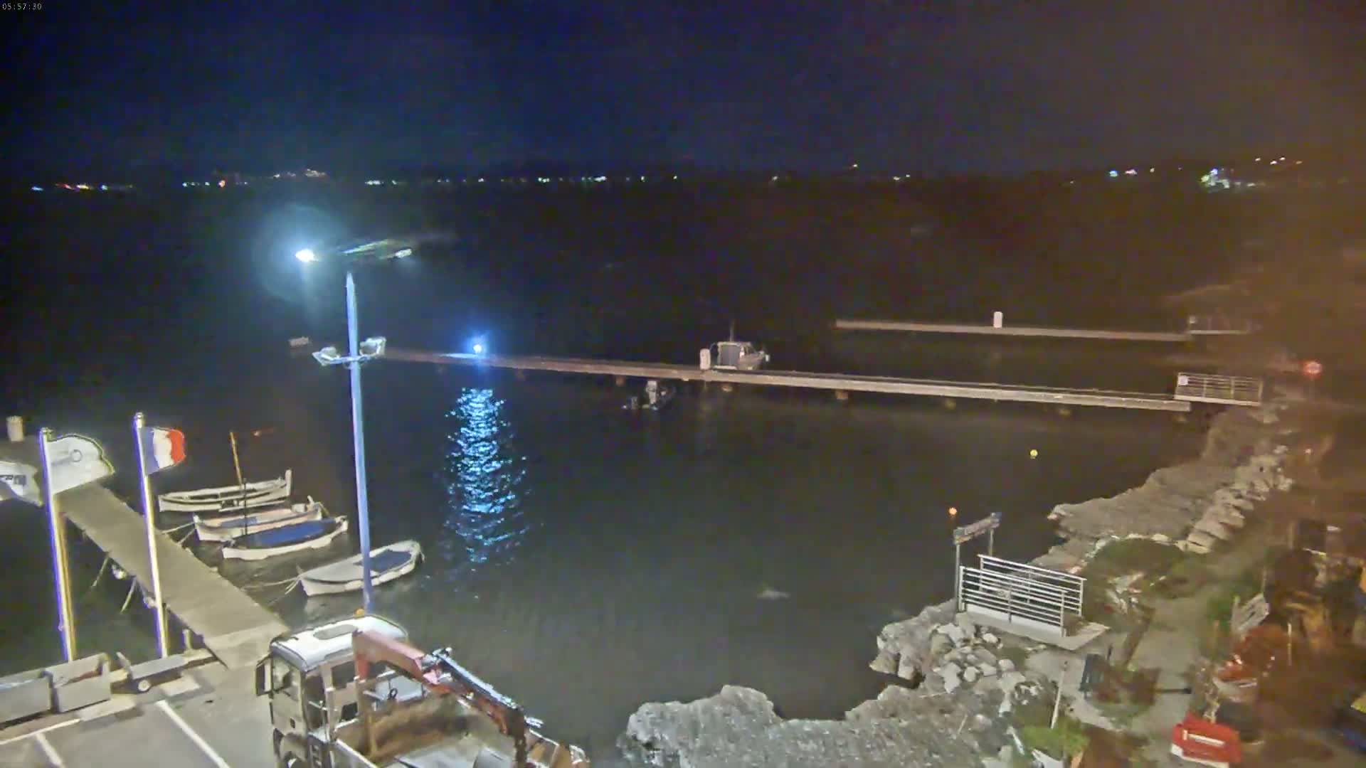 At night, several small boats are moored at a pier on a dark body of water, with a truck parked nearby and lights illuminating the scene.