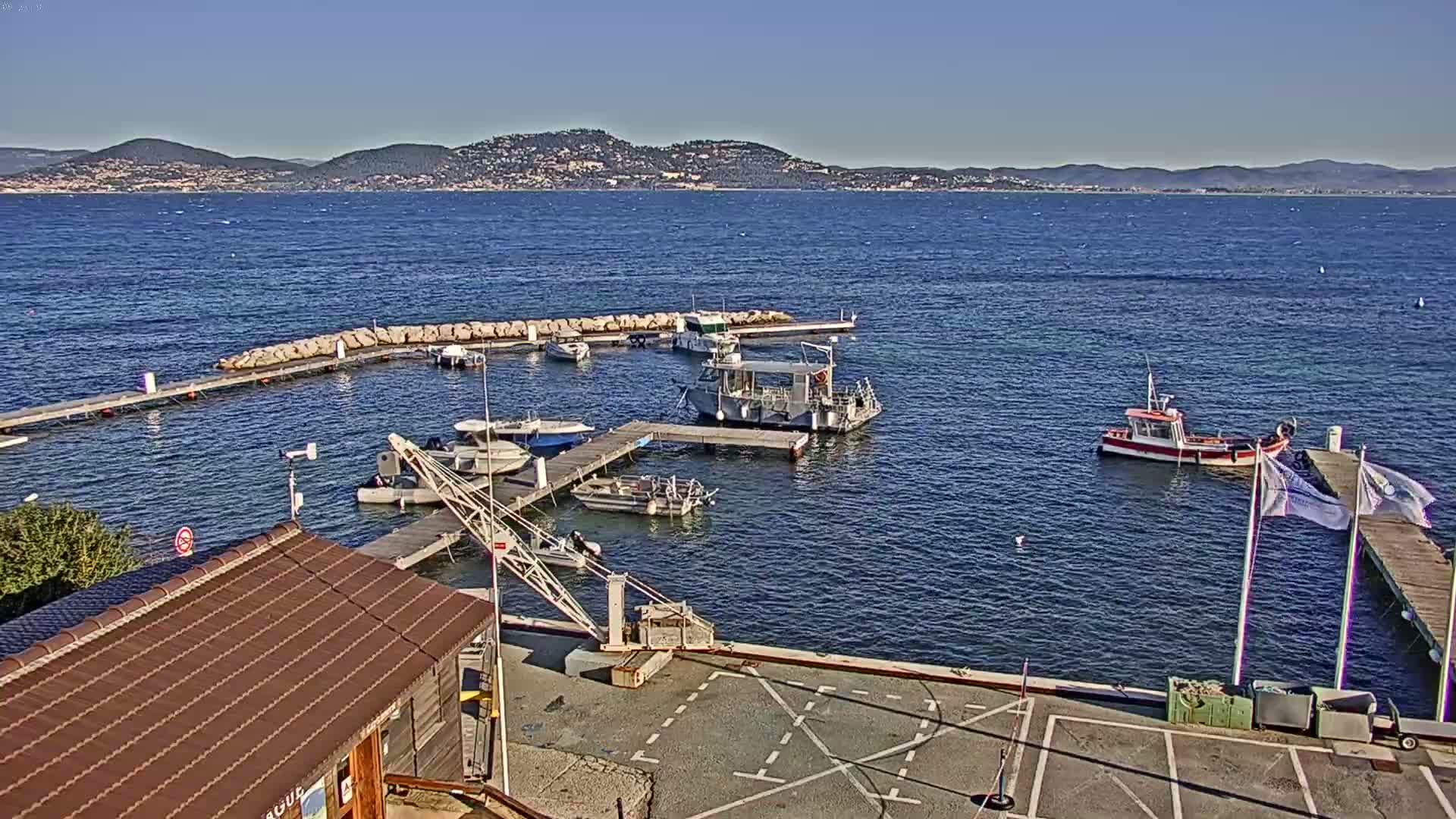 A small marina with several boats docked, a small building with a brown roof, and a clear view of a distant mountain range under a sunny sky.